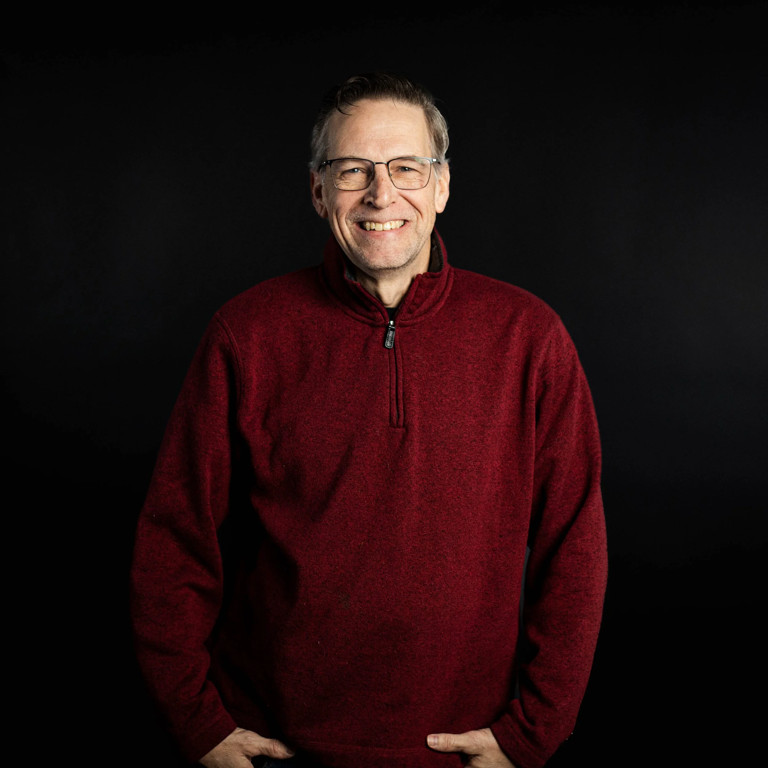 Portrait of a smiling middle-aged man wearing glasses and a red quarter-zip sweater, standing against a black background.