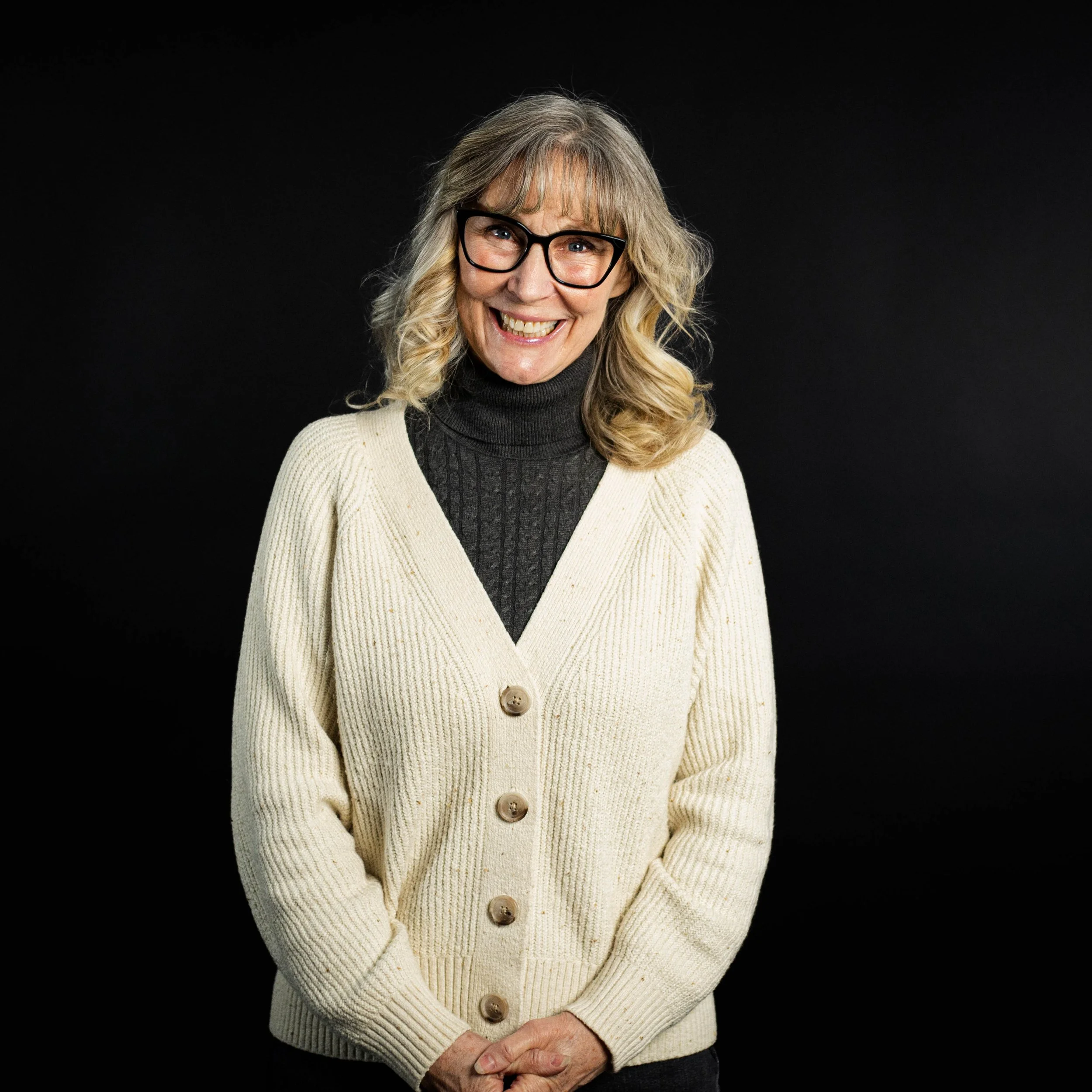 A woman with curly blonde hair and glasses smiling in front of a black background, wearing a cream-colored cardigan over a black turtleneck.