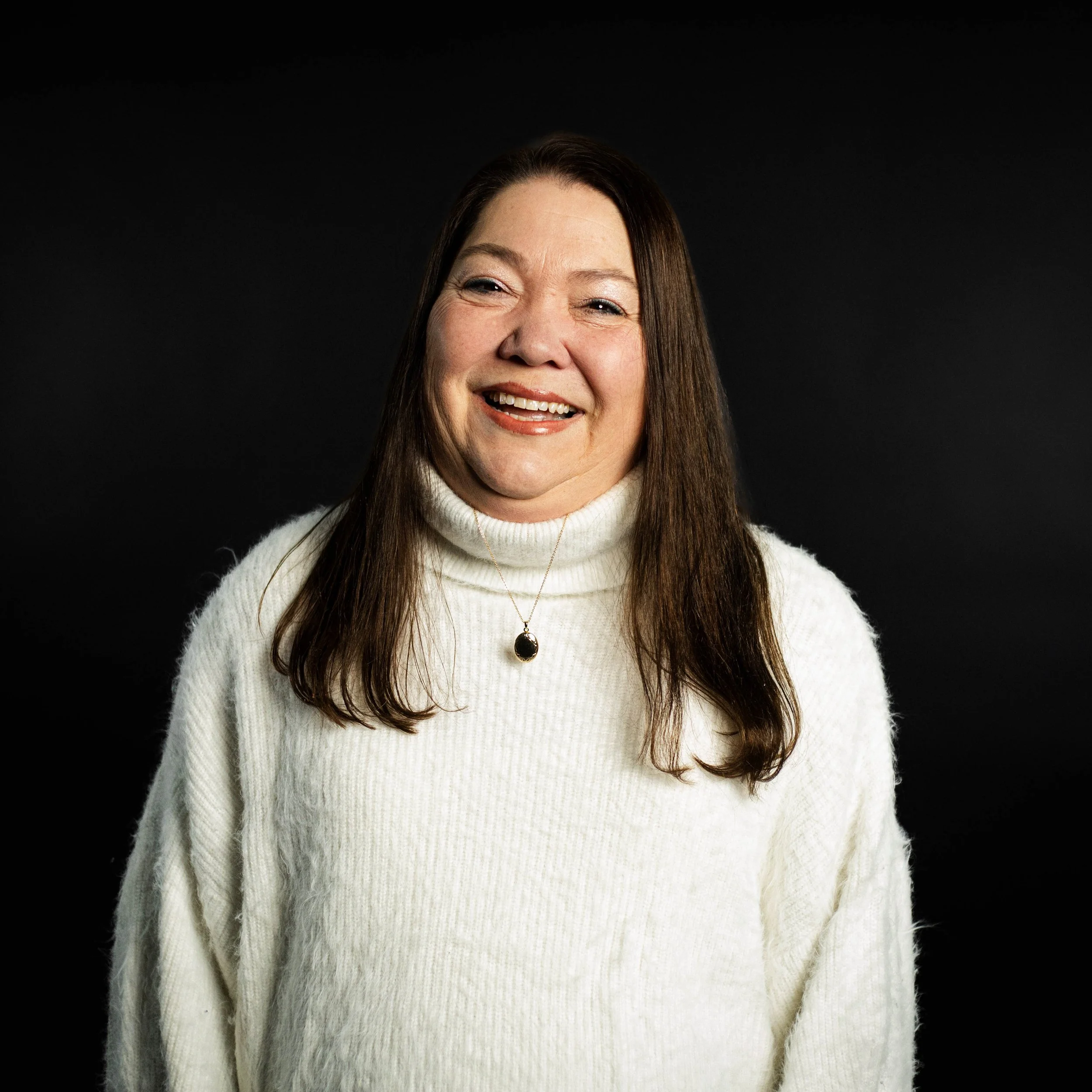 A woman with long dark hair, smiling and standing with hands on hips, wearing a beige blouse with ruffled front, against a black background.