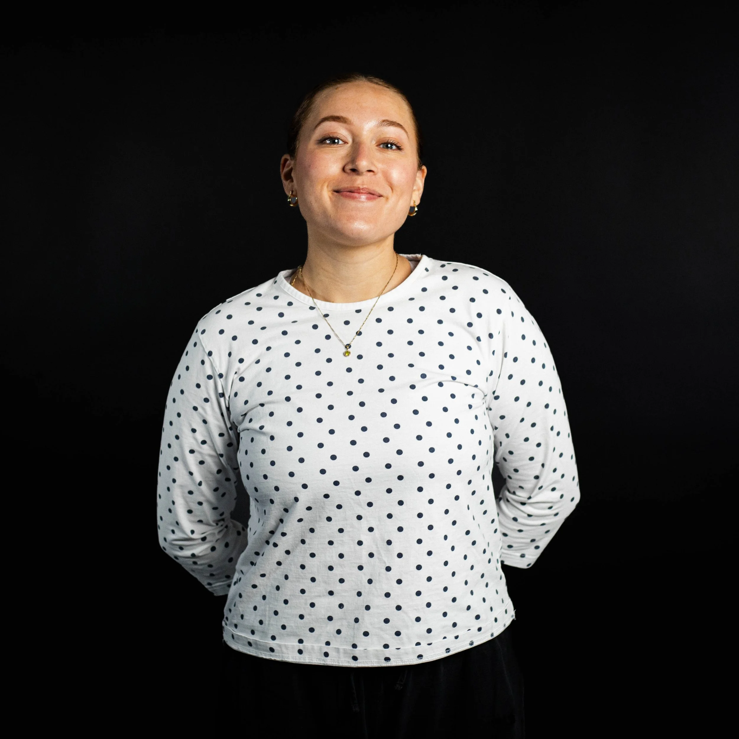 A young woman standing with her hands behind her back, smiling at the camera, wearing a white long-sleeved shirt with black polka dots and gold jewelry, against a black background.