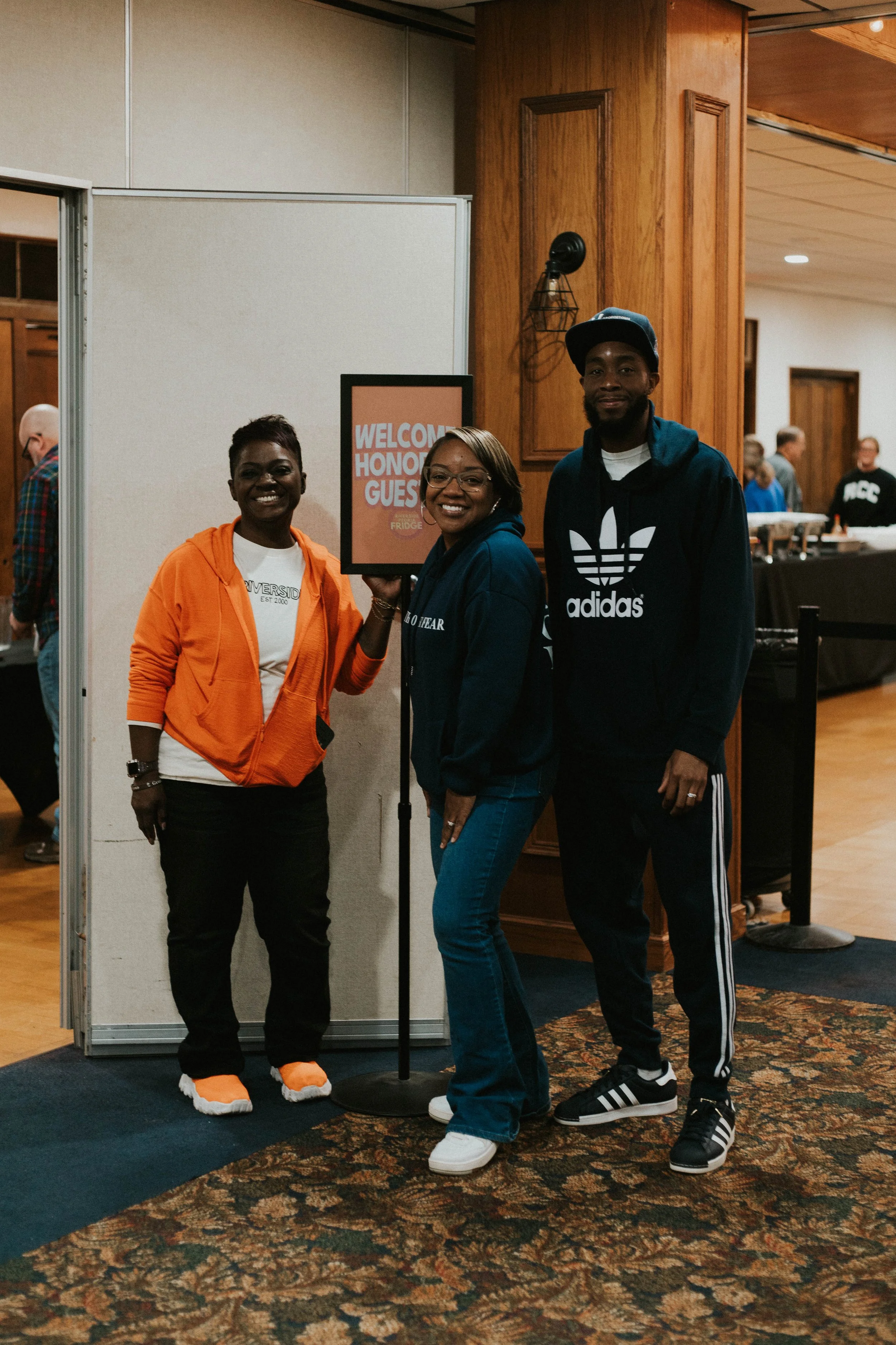 Three people standing together at a welcoming event, smiling, with a sign that says 'Welcome Honor Guests' behind them.