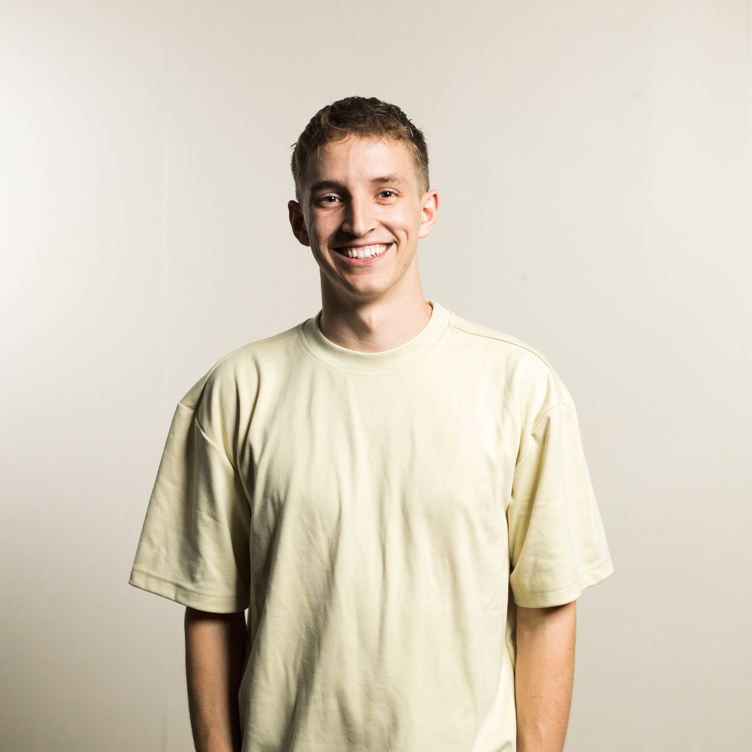 A smiling young man in a teal jacket and khaki pants standing outdoors on a grassy hill with rolling green fields in the background.