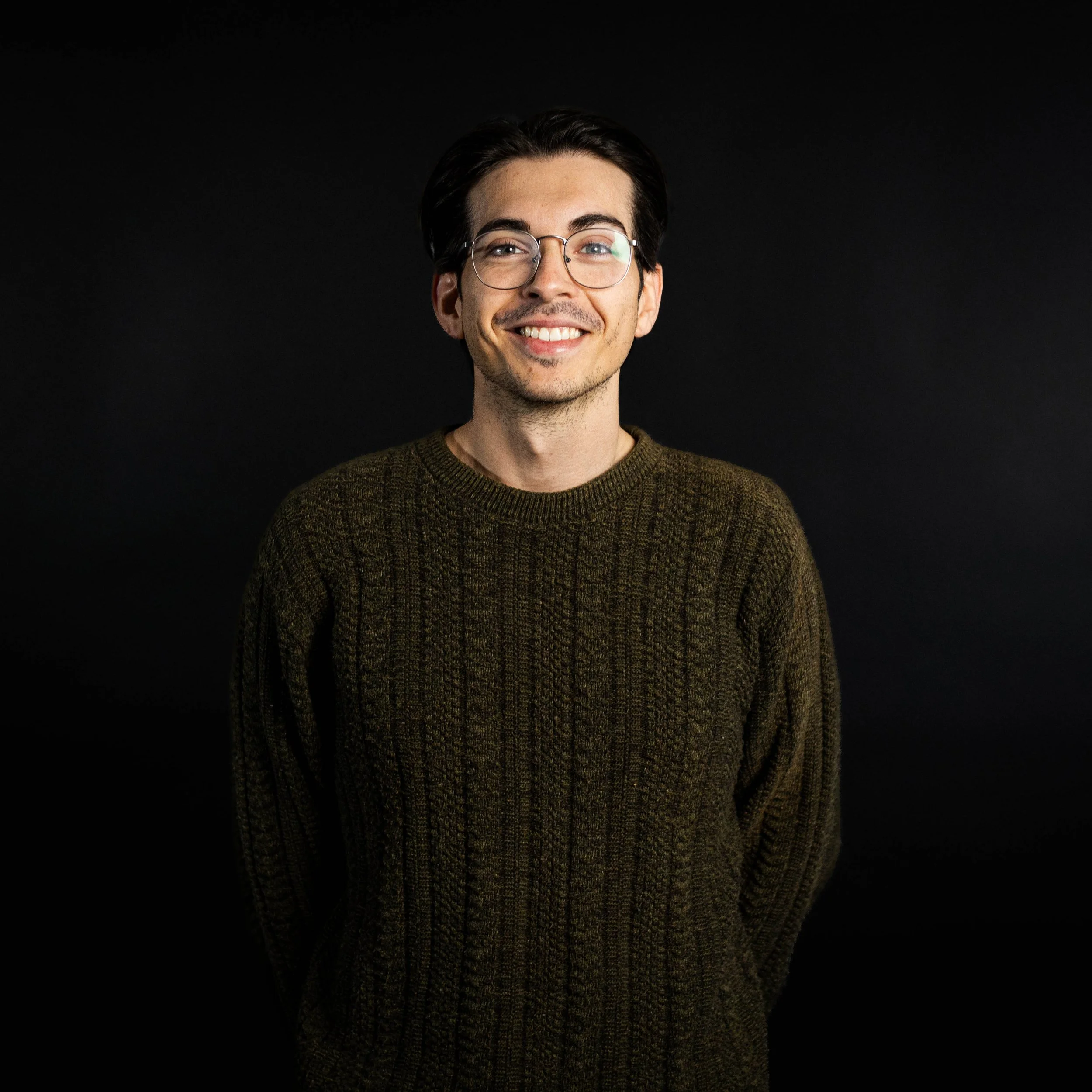 A young man with glasses, smiling, wearing a dark knit sweater, standing against a black background.