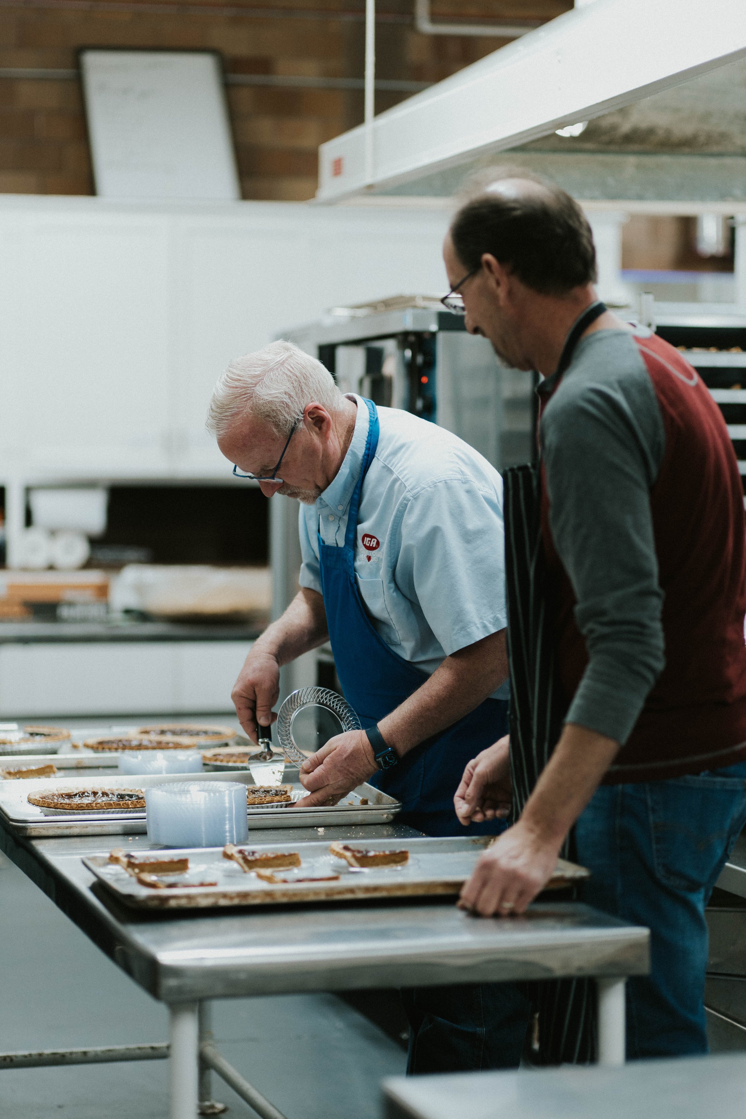 Two men working in a kitchen preparing slices of pie on trays.
