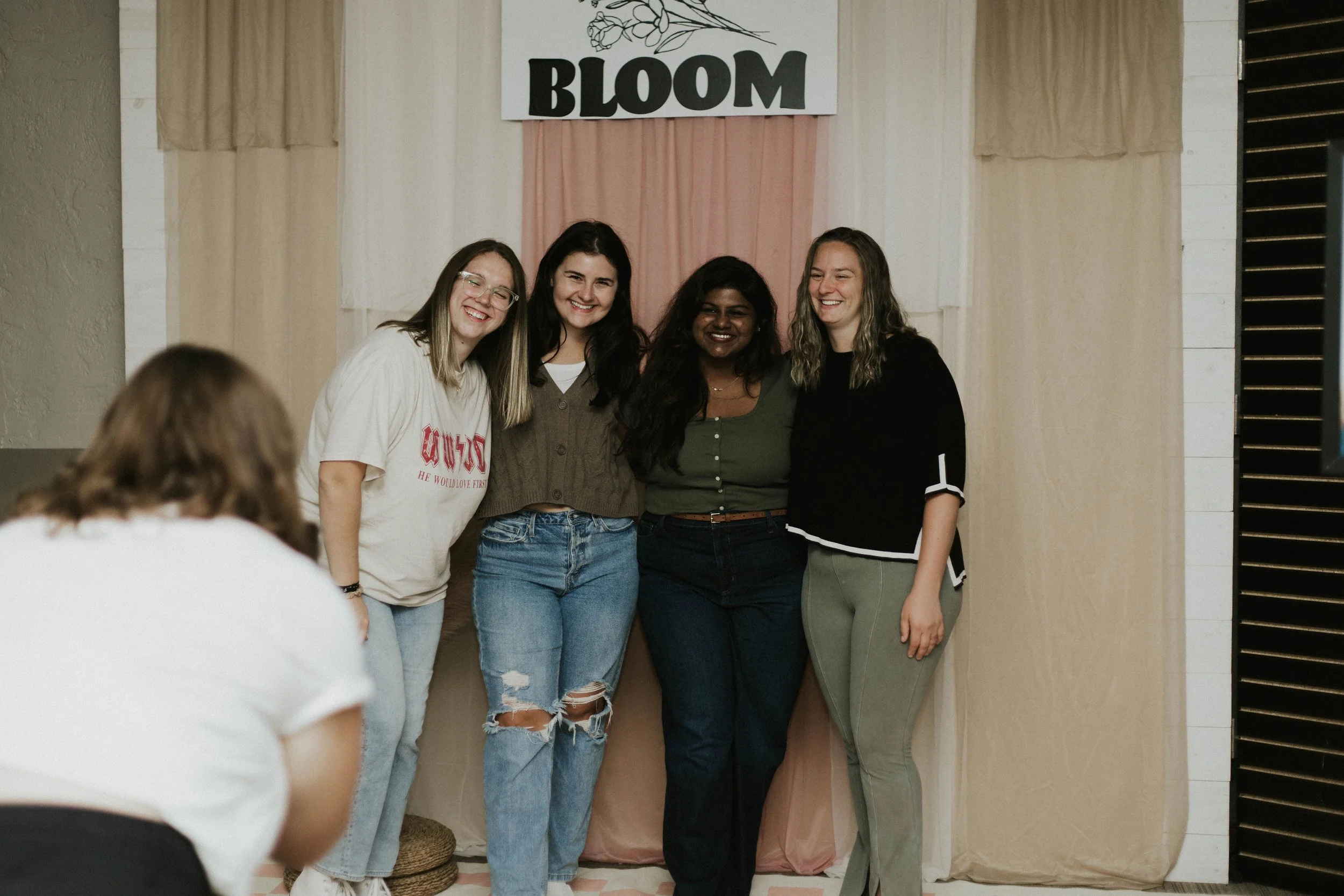 Four women standing together, smiling, in front of a backdrop with the word 'BLOOM' at a social or event setting.