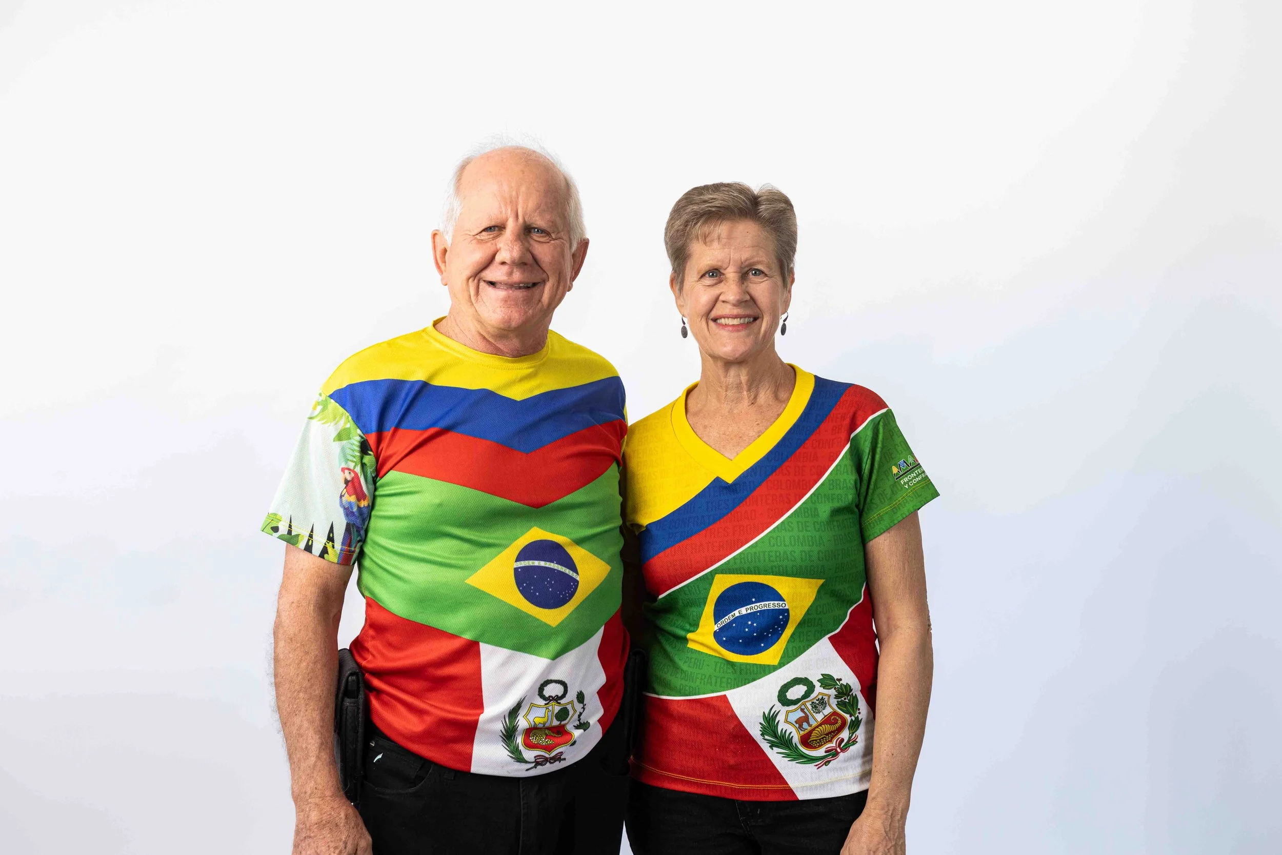 An elderly man and woman smiling and standing together, wearing colorful shirts with Brazilian, Colombian, and Peruvian flags on them, against a plain white background.