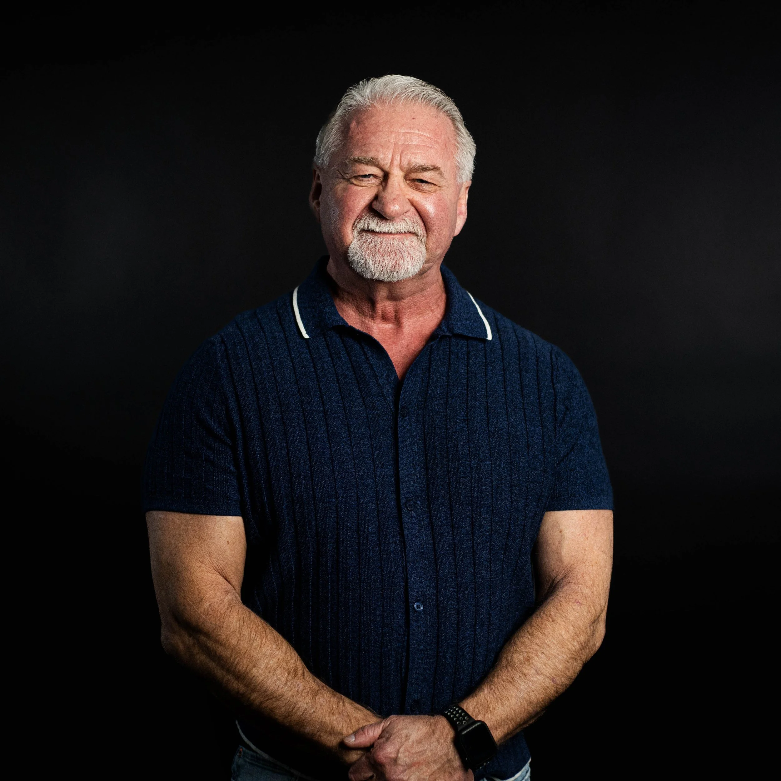 A smiling Black man wearing a denim jacket over a black t-shirt, standing against a dark plain background.
