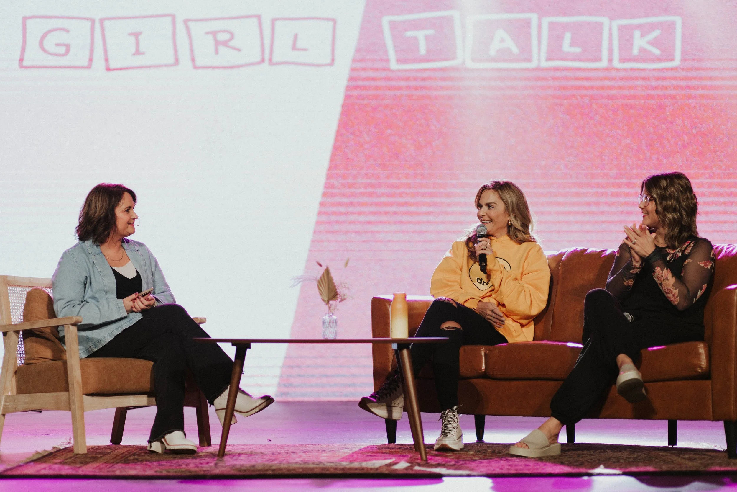 Three women sitting on sofas engaging in a discussion on stage with a backdrop that reads 'GIRL TALK'.