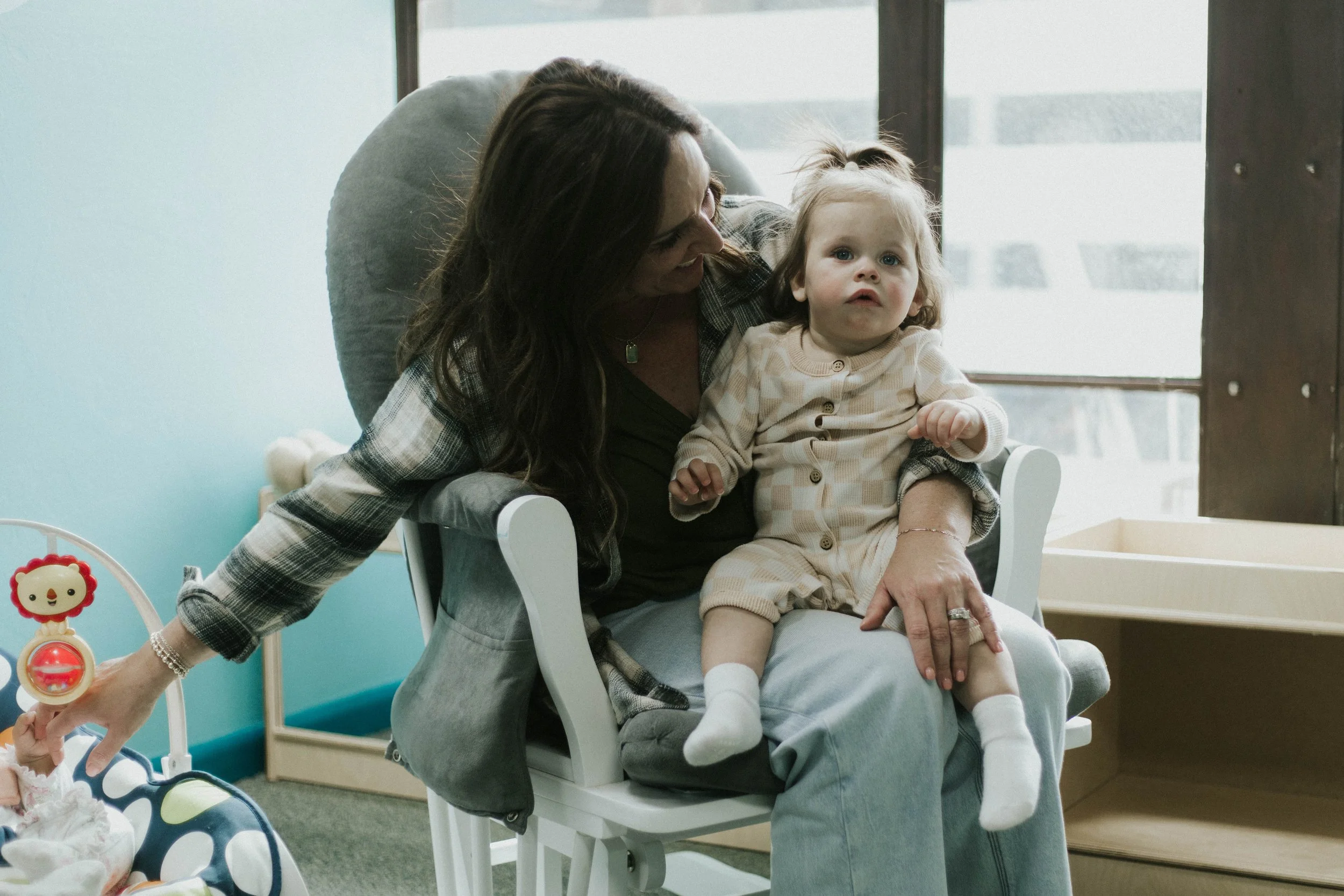 A woman sitting on a chair holding a young girl on her lap, both are indoors near a window. The woman is leaning towards the girl, and they are both looking in the same direction. The girl has a small ponytail and is wearing a beige plaid outfit.