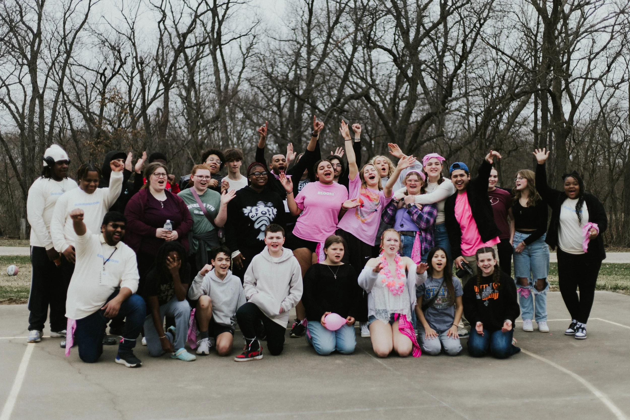 A large group of diverse young people gathered outdoors on a gray day, raising their hands and smiling, on a paved court with leafless trees in the background, celebrating or participating in a group activity.