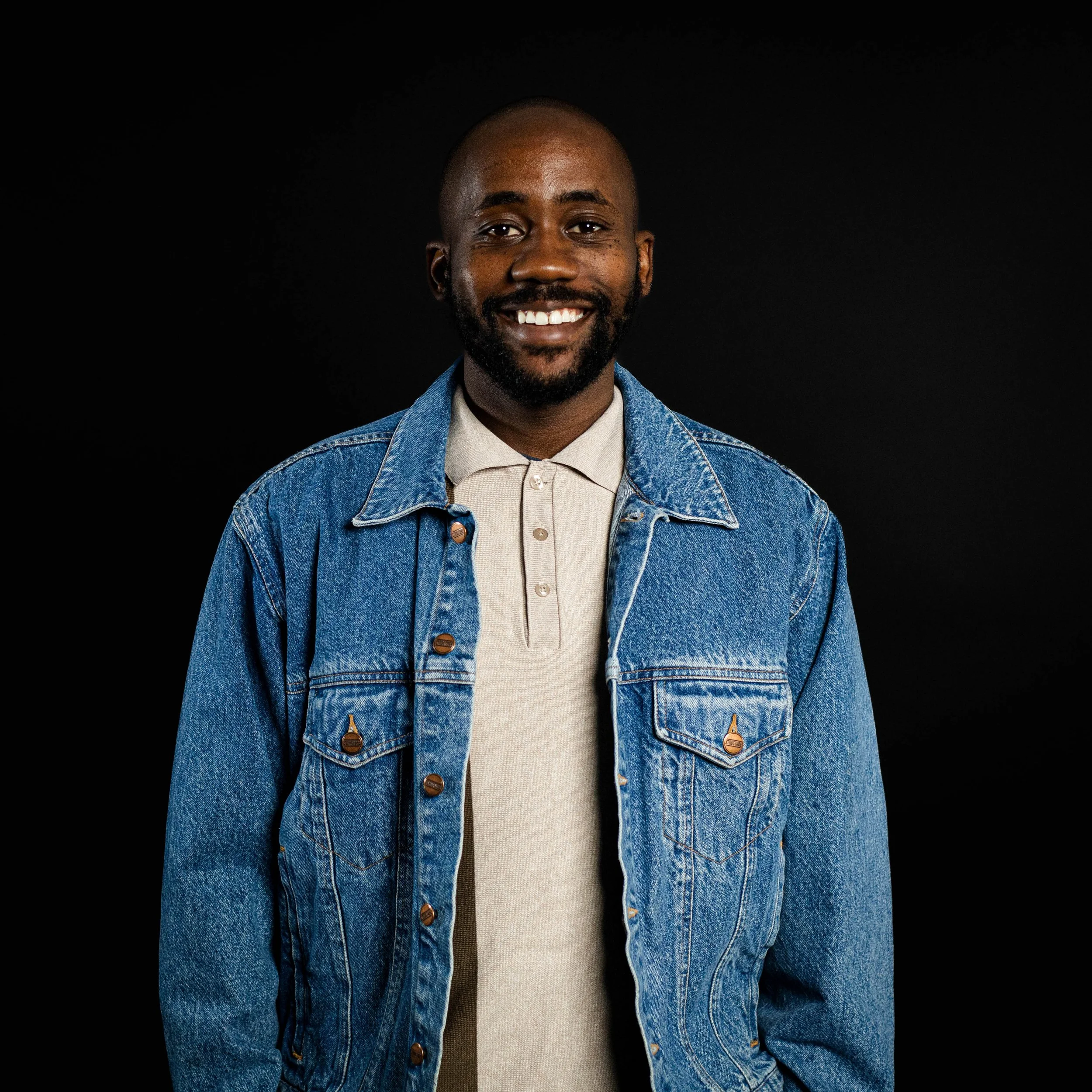 A smiling Black man wearing a denim jacket over a black t-shirt, standing against a dark plain background.