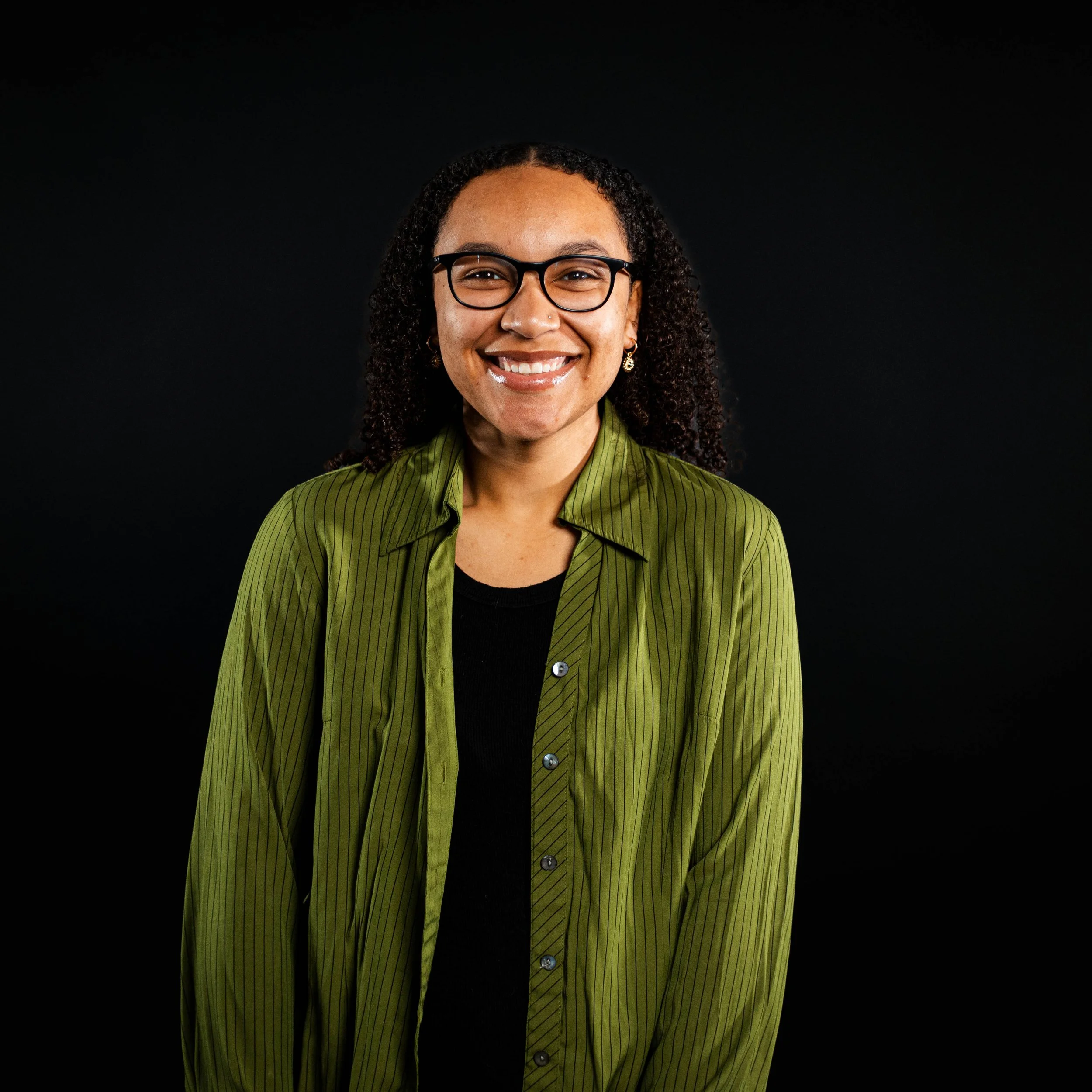 Smiling woman with glasses and earrings wearing a green striped shirt against a black background.