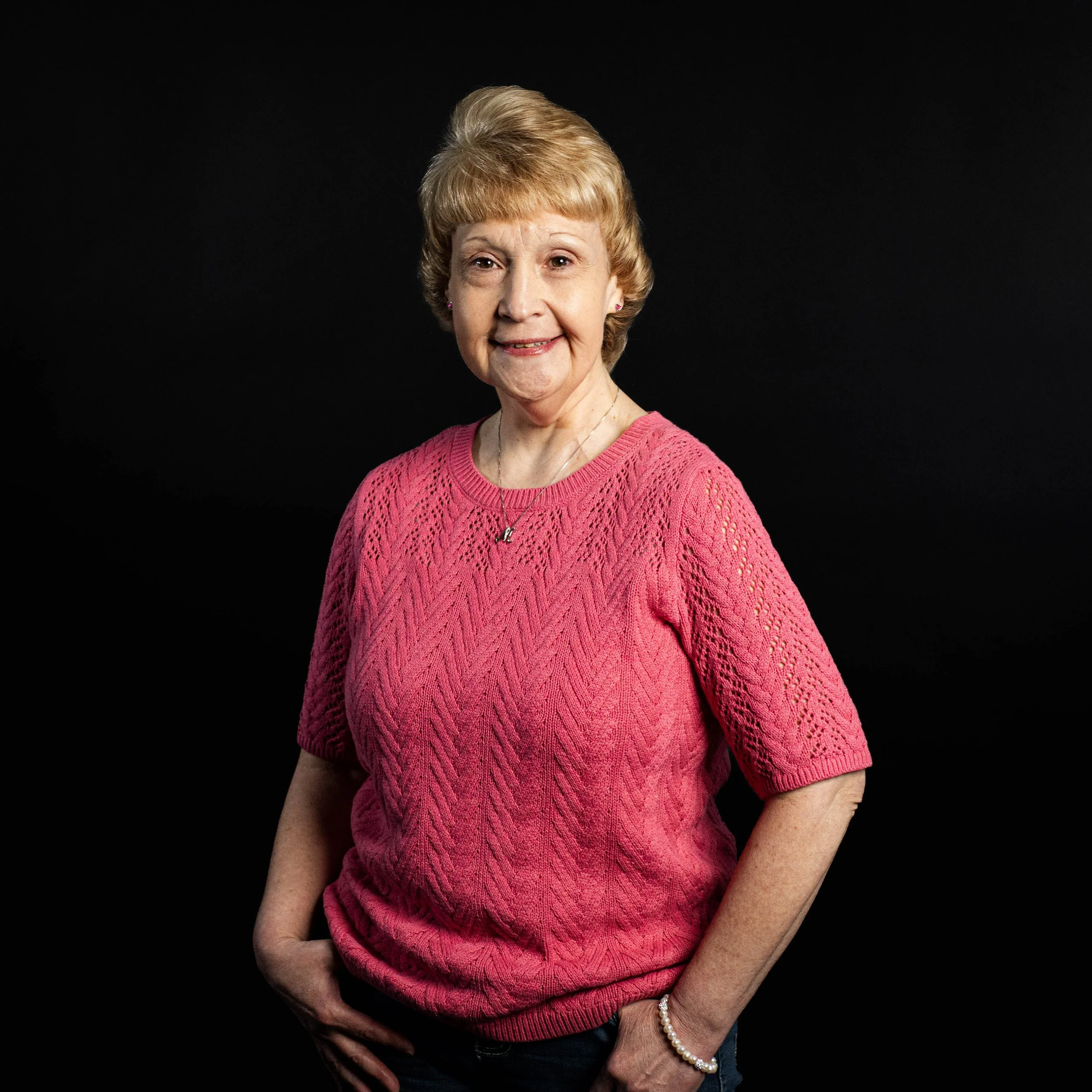 A smiling elderly woman with short blonde hair wearing a pink knitted sweater, dark jeans, a pearl bracelet, and a necklace, standing against a black background.