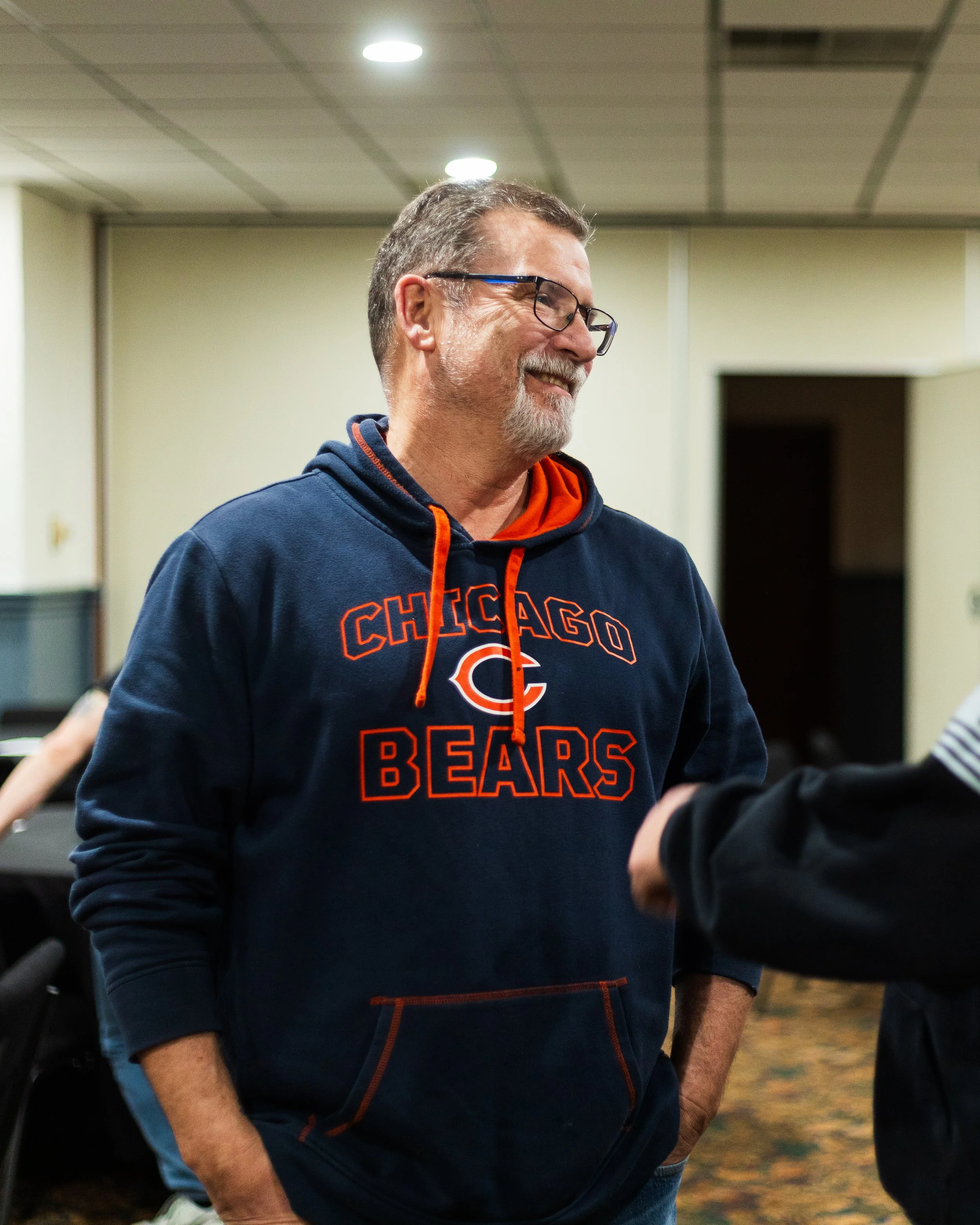 A smiling man in a navy blue Chicago Bears hoodie and glasses standing indoors, engaged in conversation with someone off-camera.