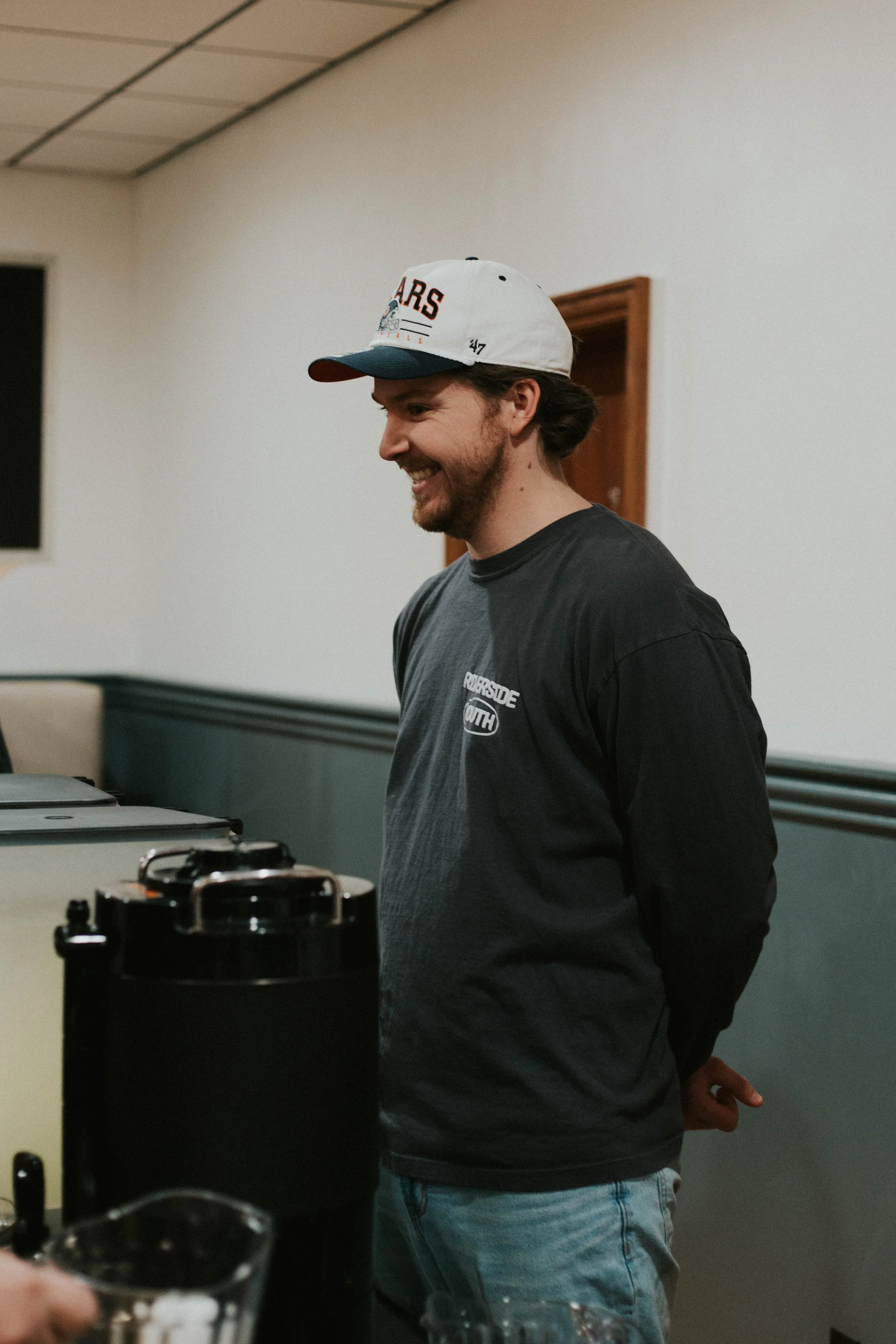A man with a beard and long hair, wearing a white and green cap and a black t-shirt, standing in profile, smiling in an indoor setting.