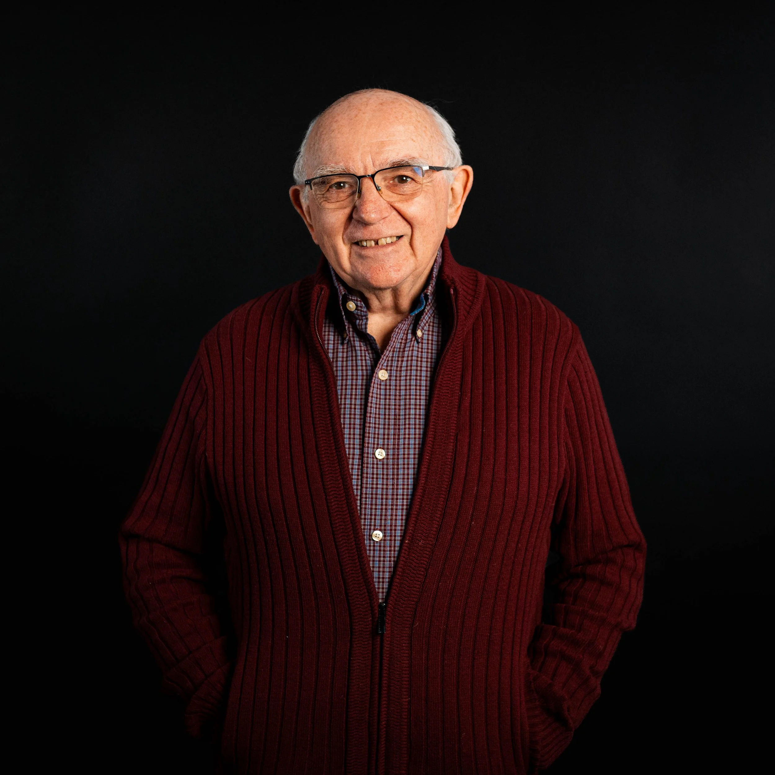An elderly man with glasses, wearing a red cardigan over a checkered shirt, standing against a black background.