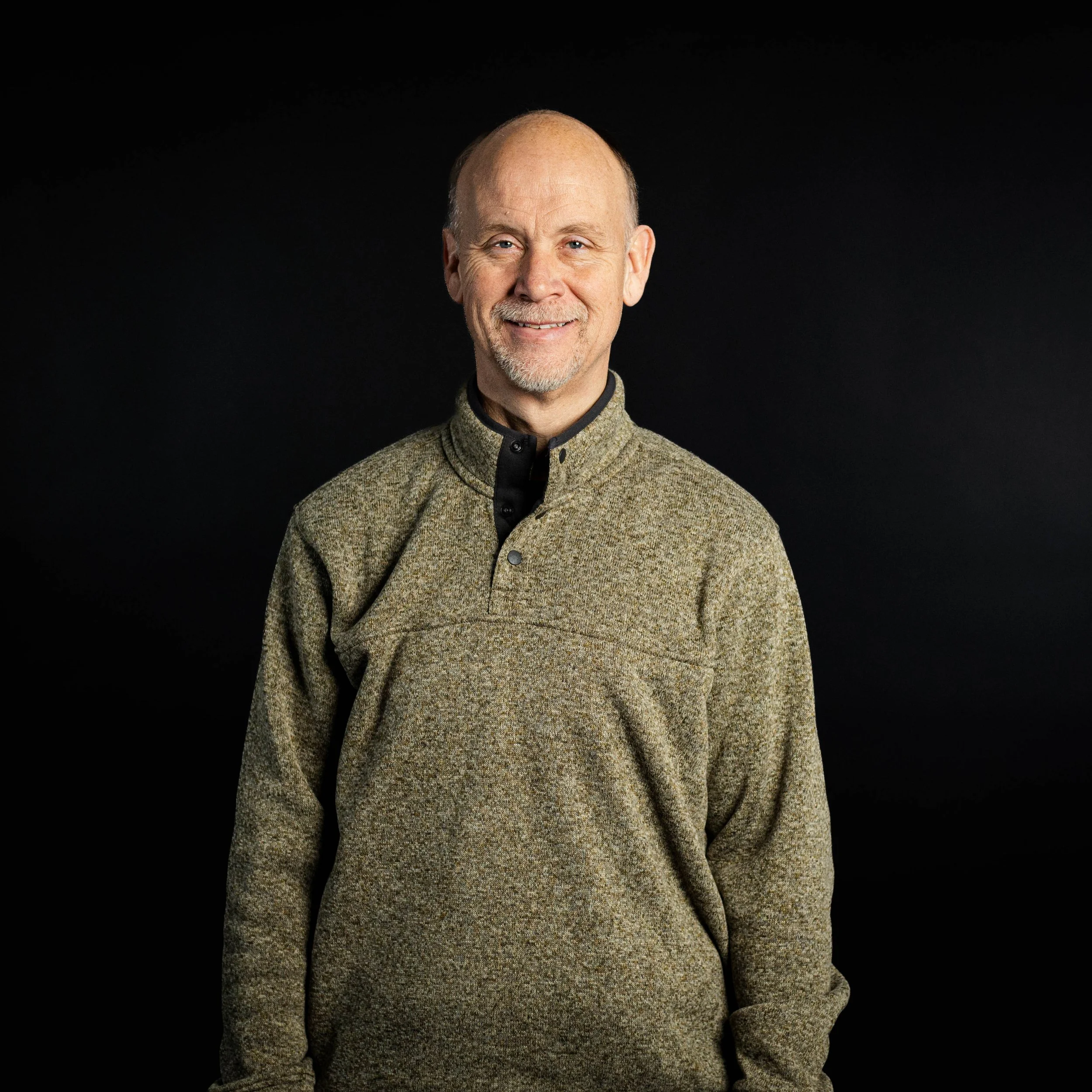 A middle-aged man with a bald head, smiling, wearing a beige and black pullover in front of a black background.