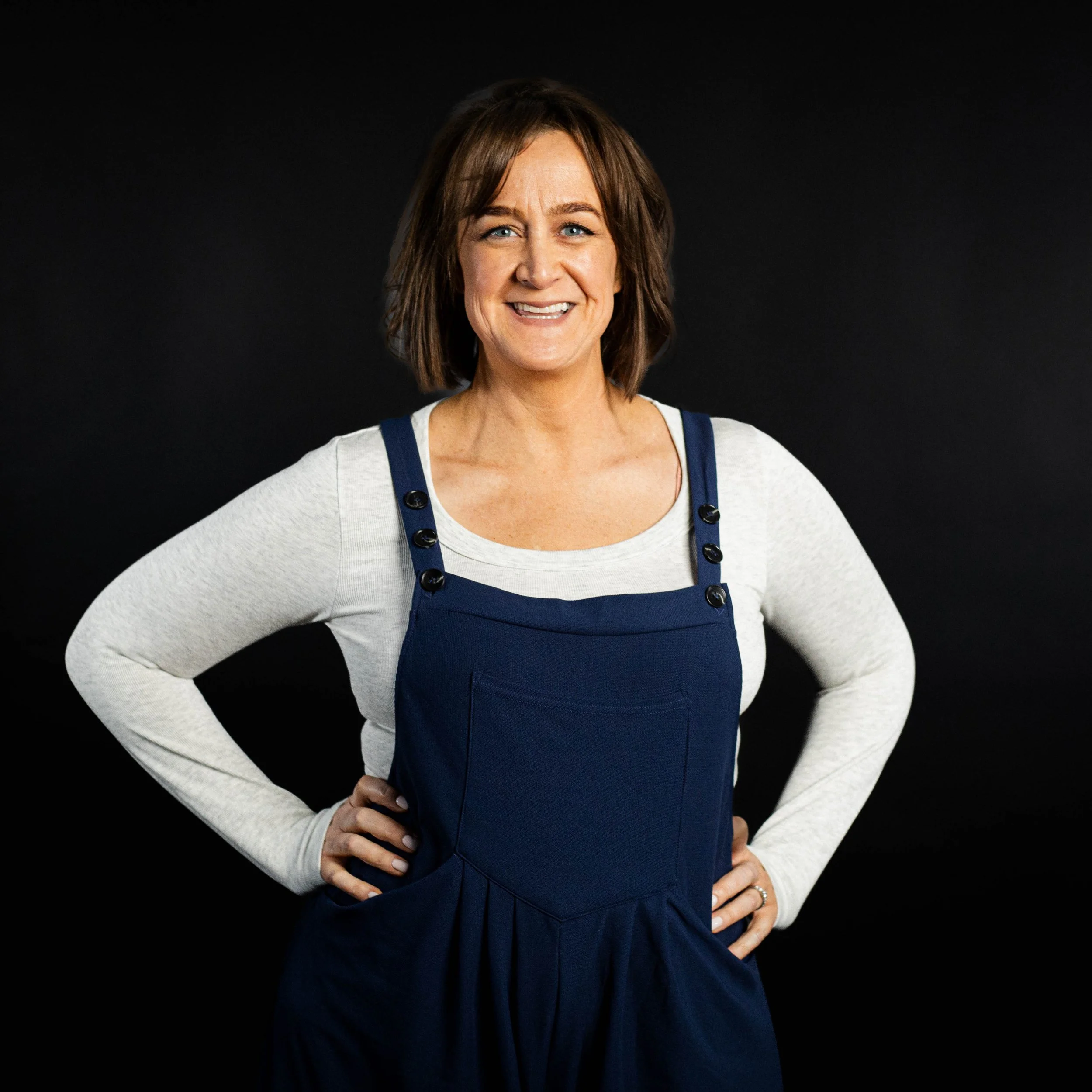 Woman with short brown hair smiling, wearing a white long-sleeve shirt and navy blue overalls with buttons, standing against a black background.
