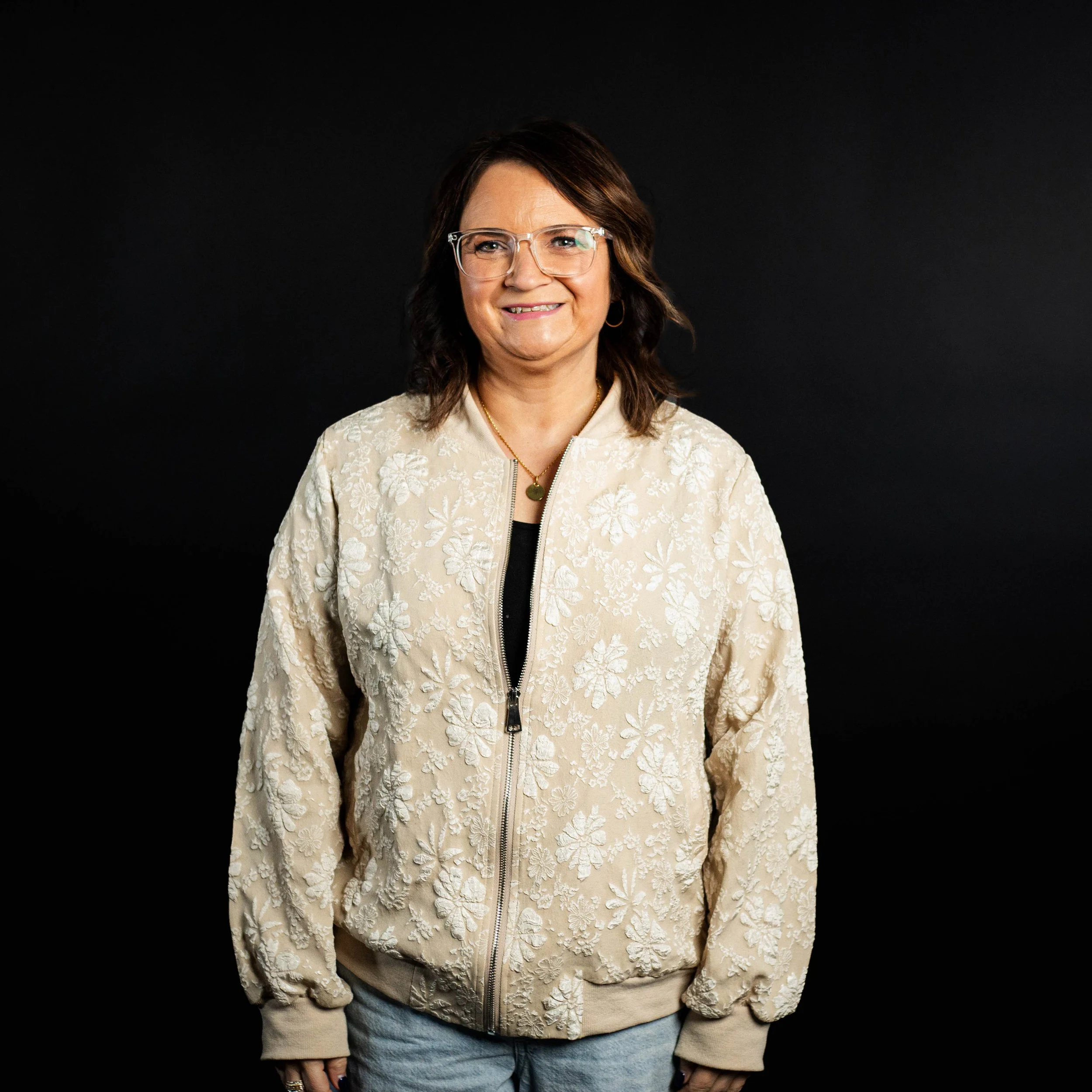 Woman with glasses smiling, wearing a cream-colored floral embroidered zip-up jacket and light blue jeans, standing against a black background.