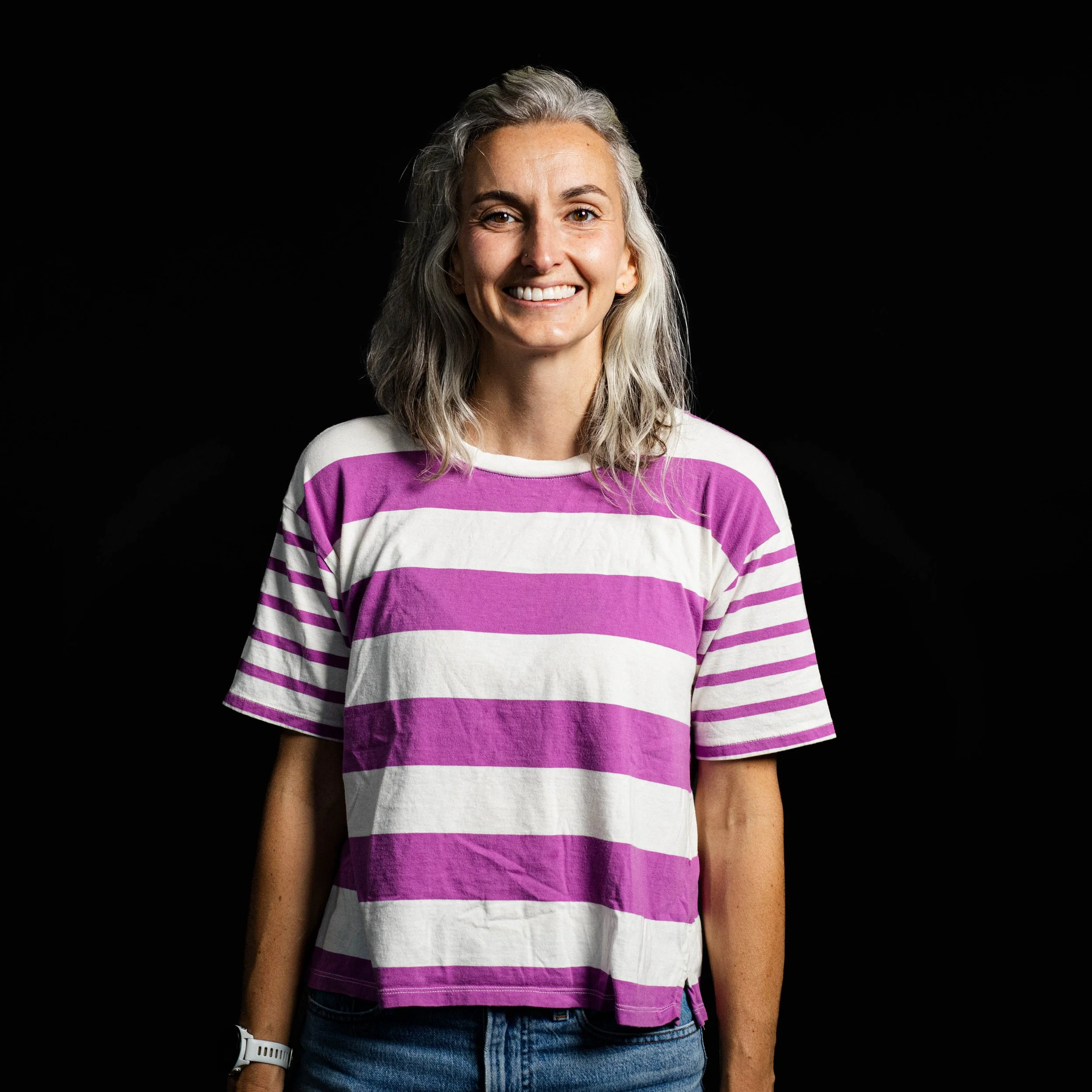 Close-up of a smiling woman with gray hair wearing a purple and white striped shirt, denim jeans, and a white watch on her wrist, standing against a black background.