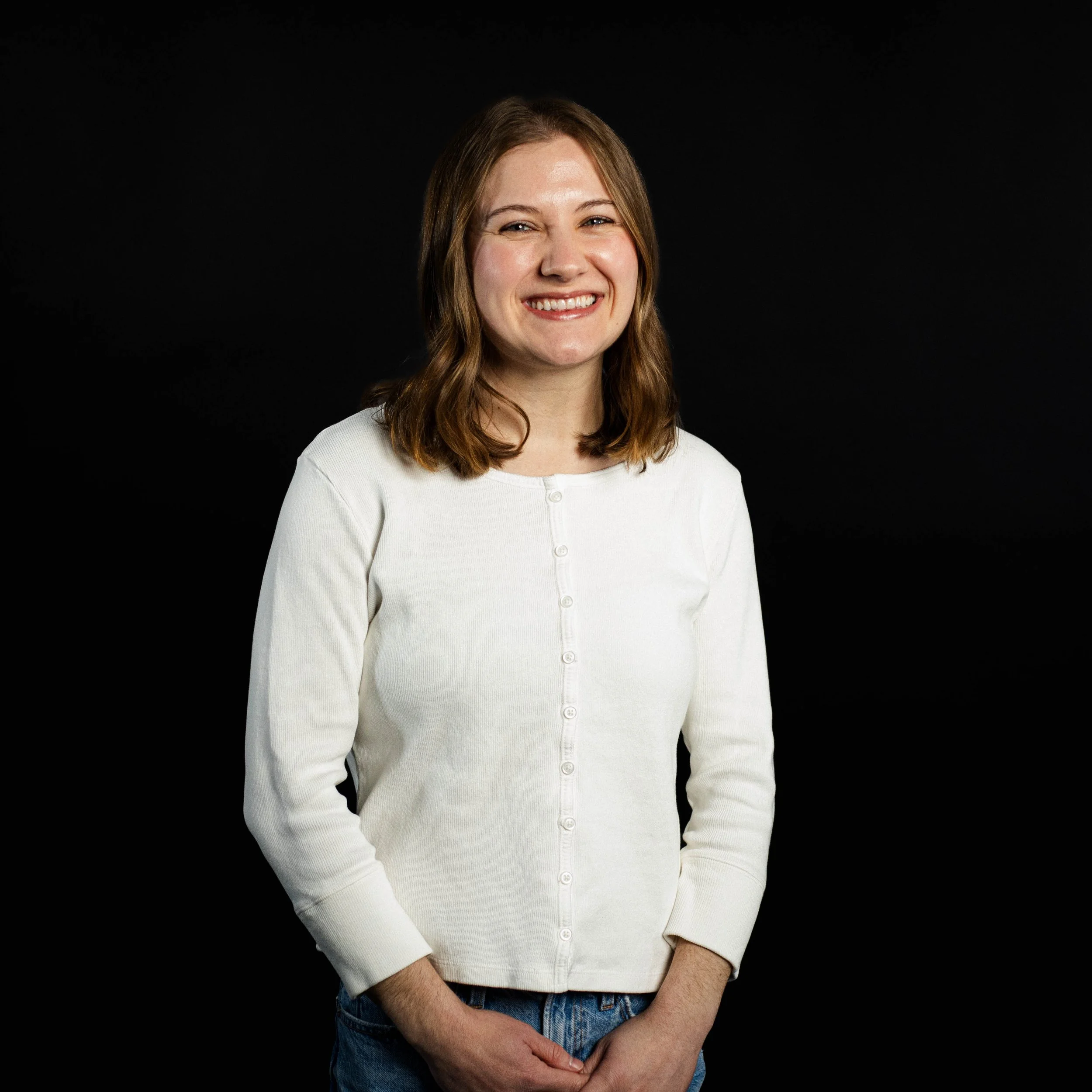 A woman with shoulder-length brown hair, smiling, wearing a cream-colored button-up sweater and blue jeans against a black background.