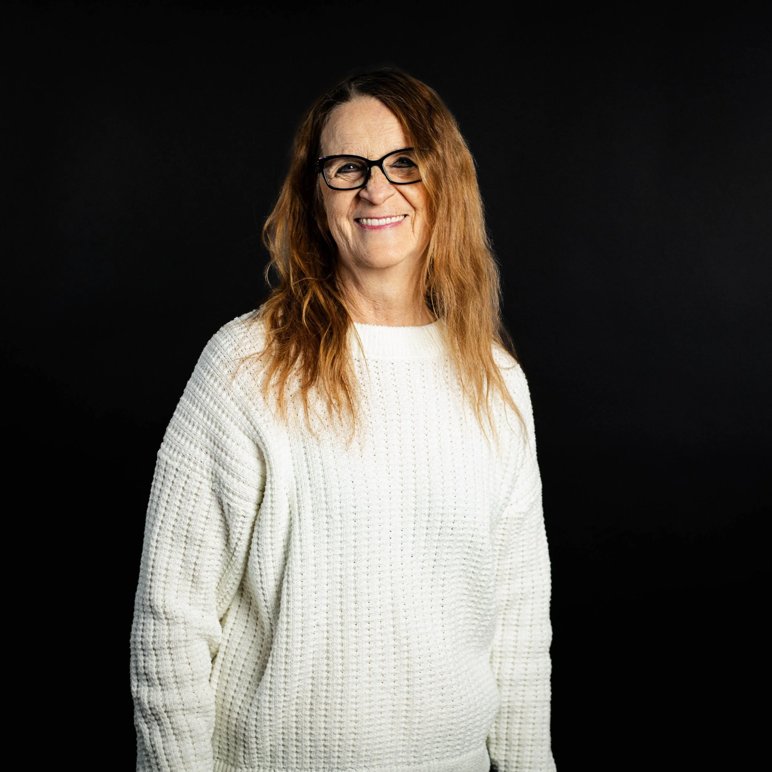 A woman with long red hair, wearing glasses and a white knitted sweater, smiling against a black background.