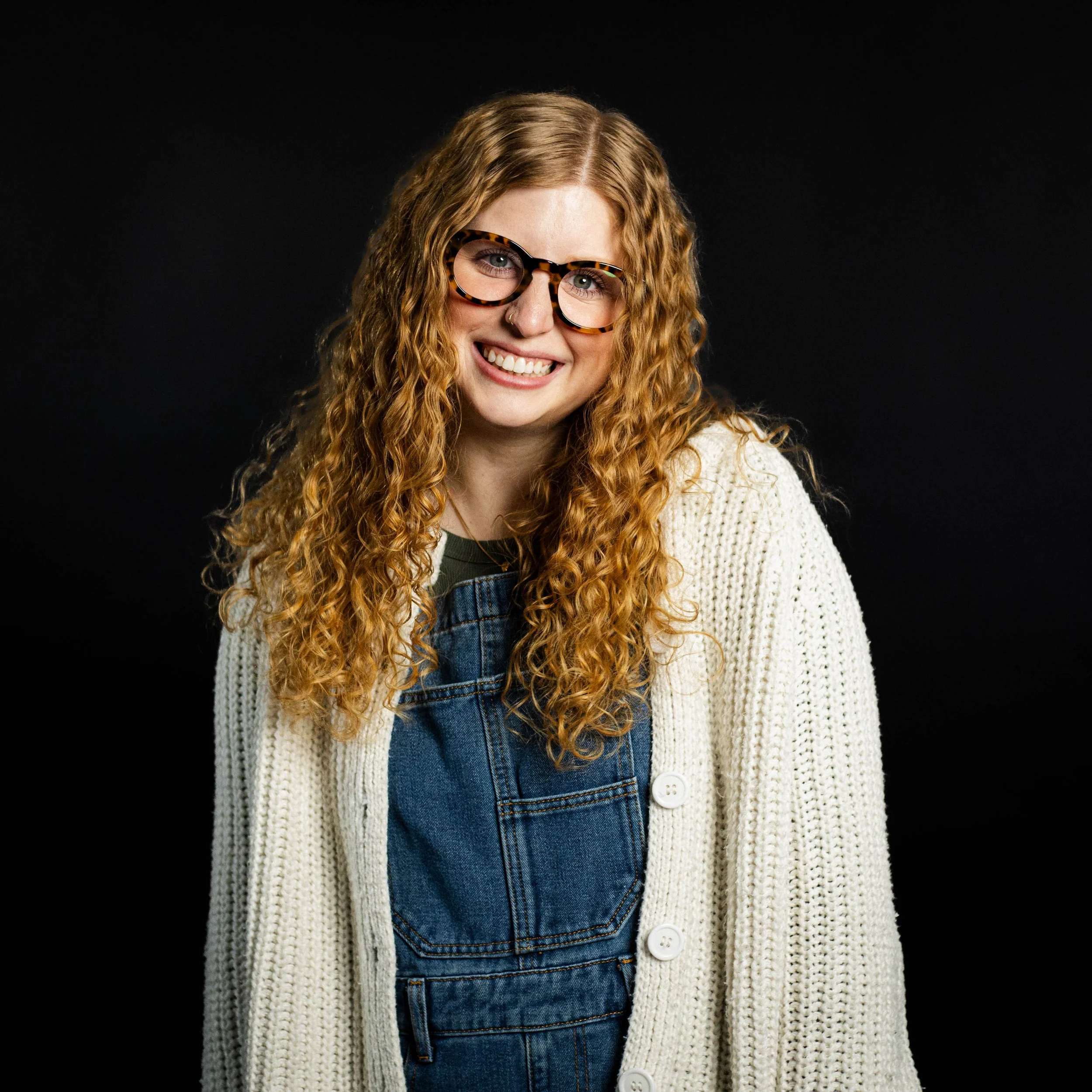 A young woman with curly red hair, wearing glasses, a white knit cardigan, and denim overalls, smiling against a black background.