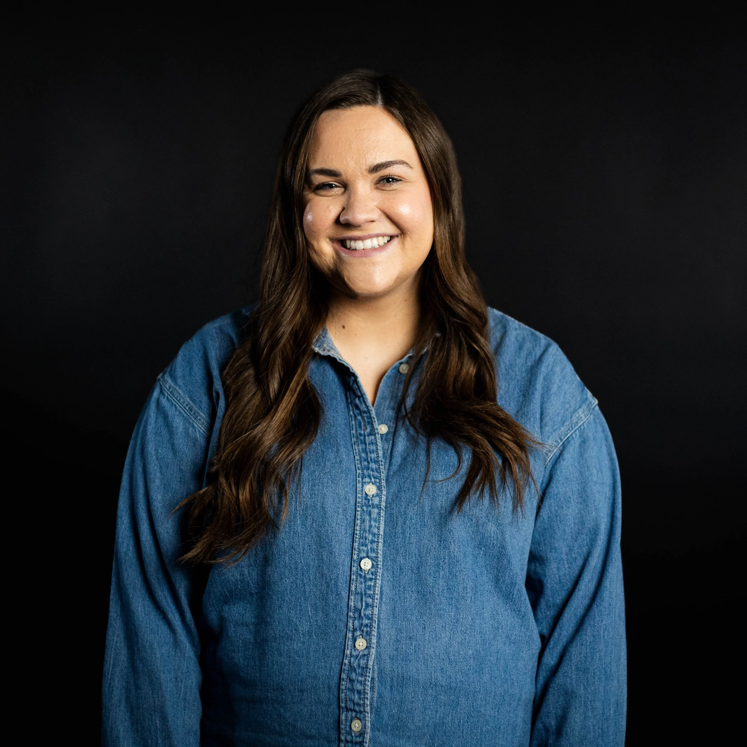 A young woman with long dark hair smiling and wearing a cream-colored embroidered blouse, standing against a dark background.