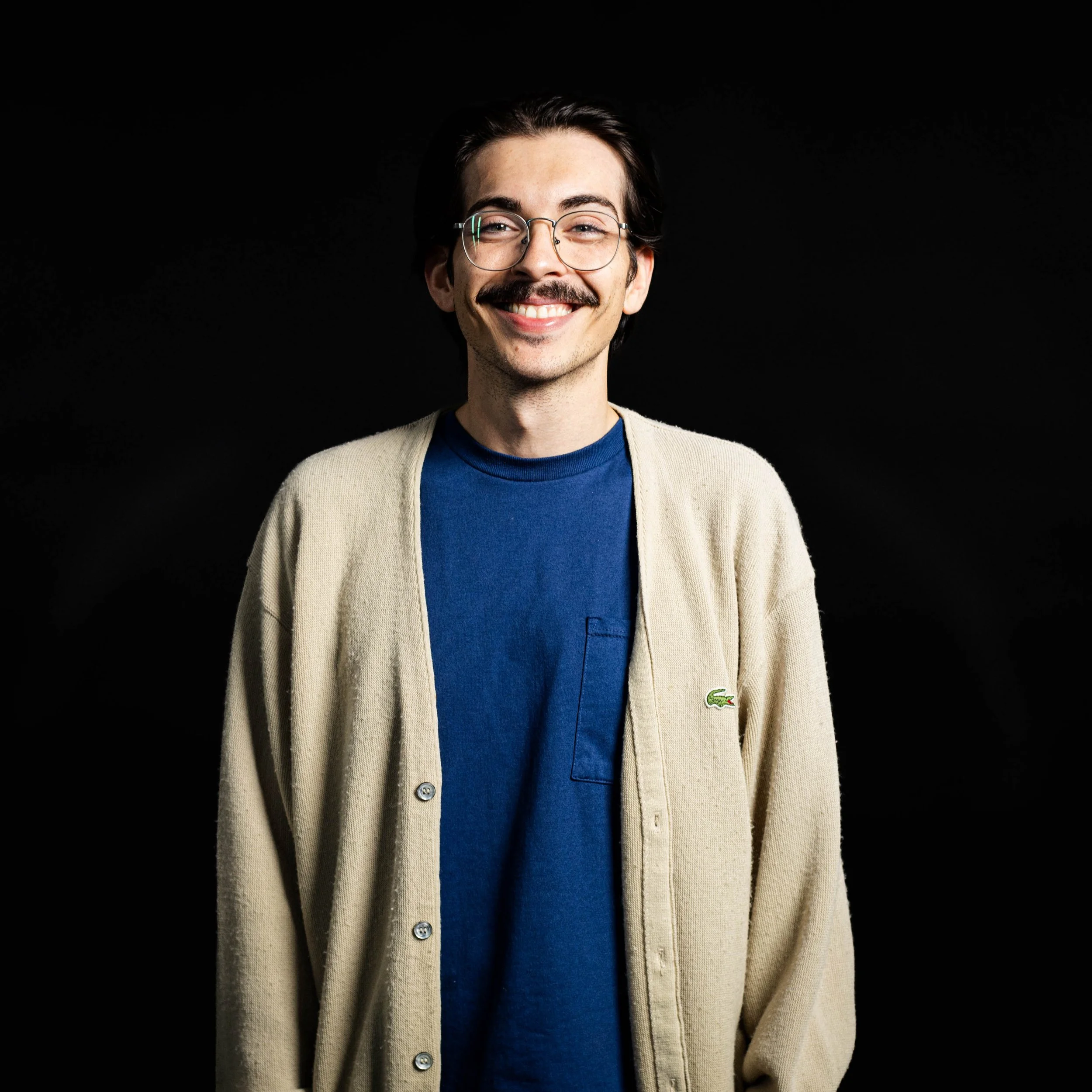 Portrait of a young man with dark hair, glasses, and a light beard, smiling, wearing a green knit sweater, standing against a black background.
