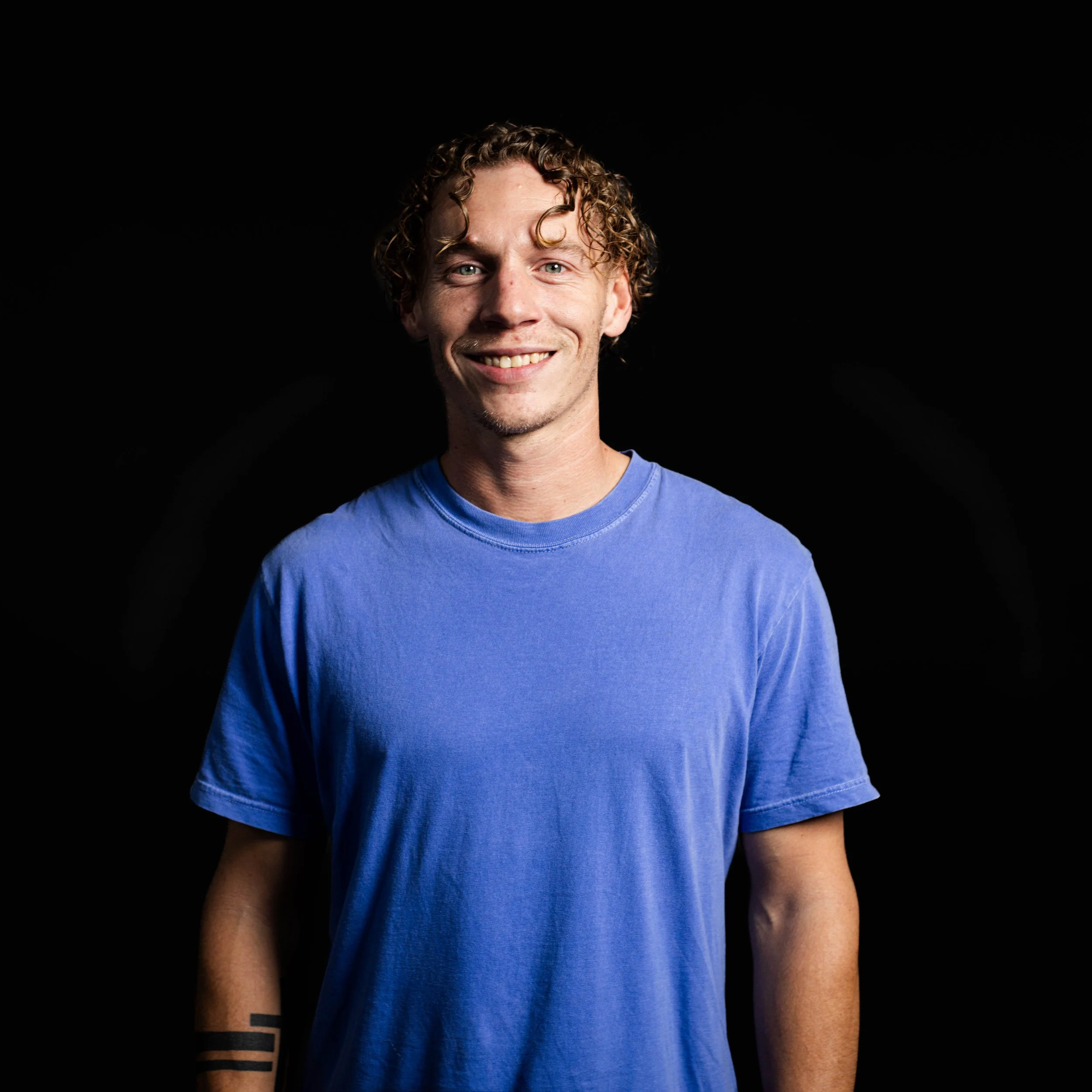 A young man with curly blond hair, blue eyes, and a light beard, smiling, wearing a blue t-shirt, against a black background.