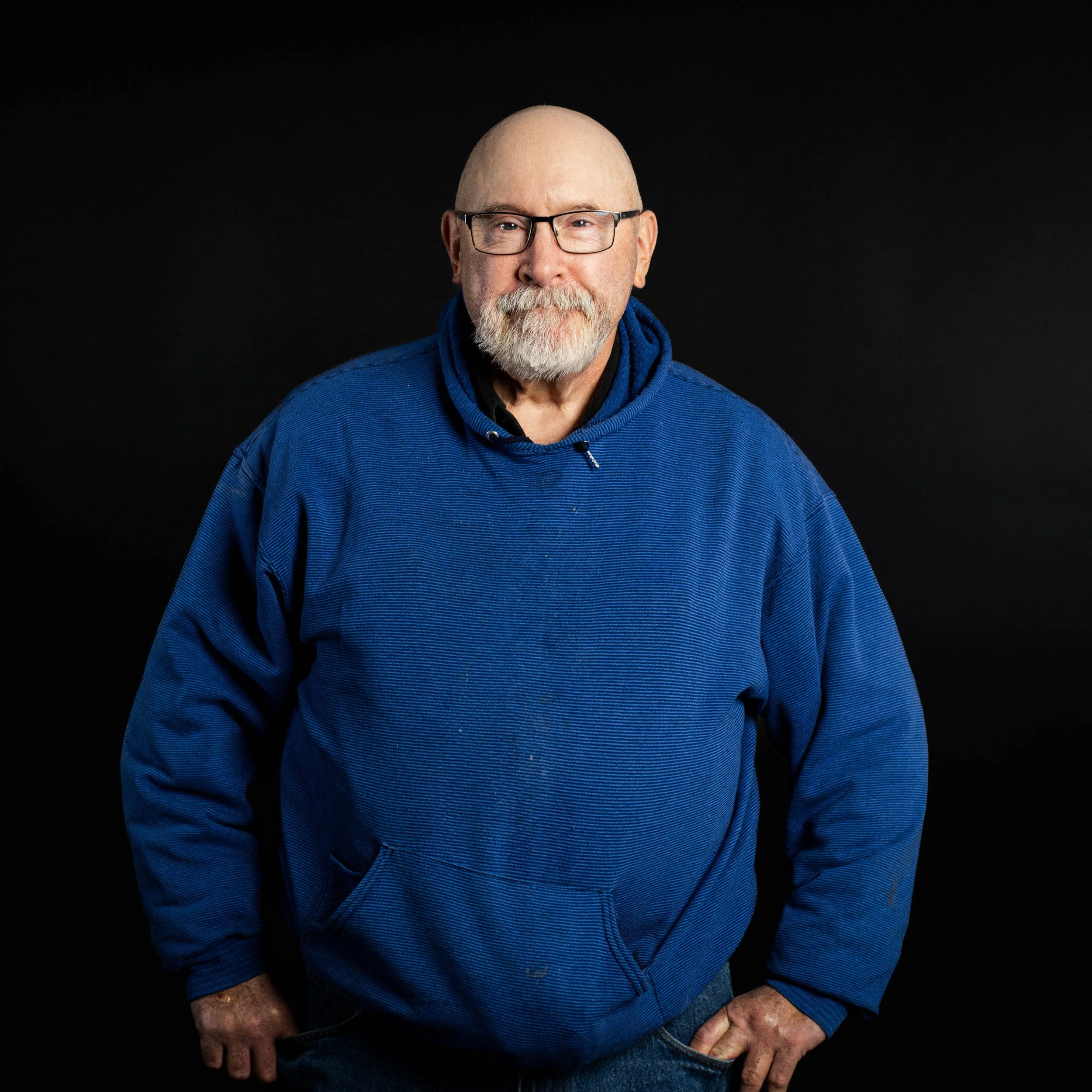 An older man with a white beard and glasses posing against a black background, wearing a blue hoodie.