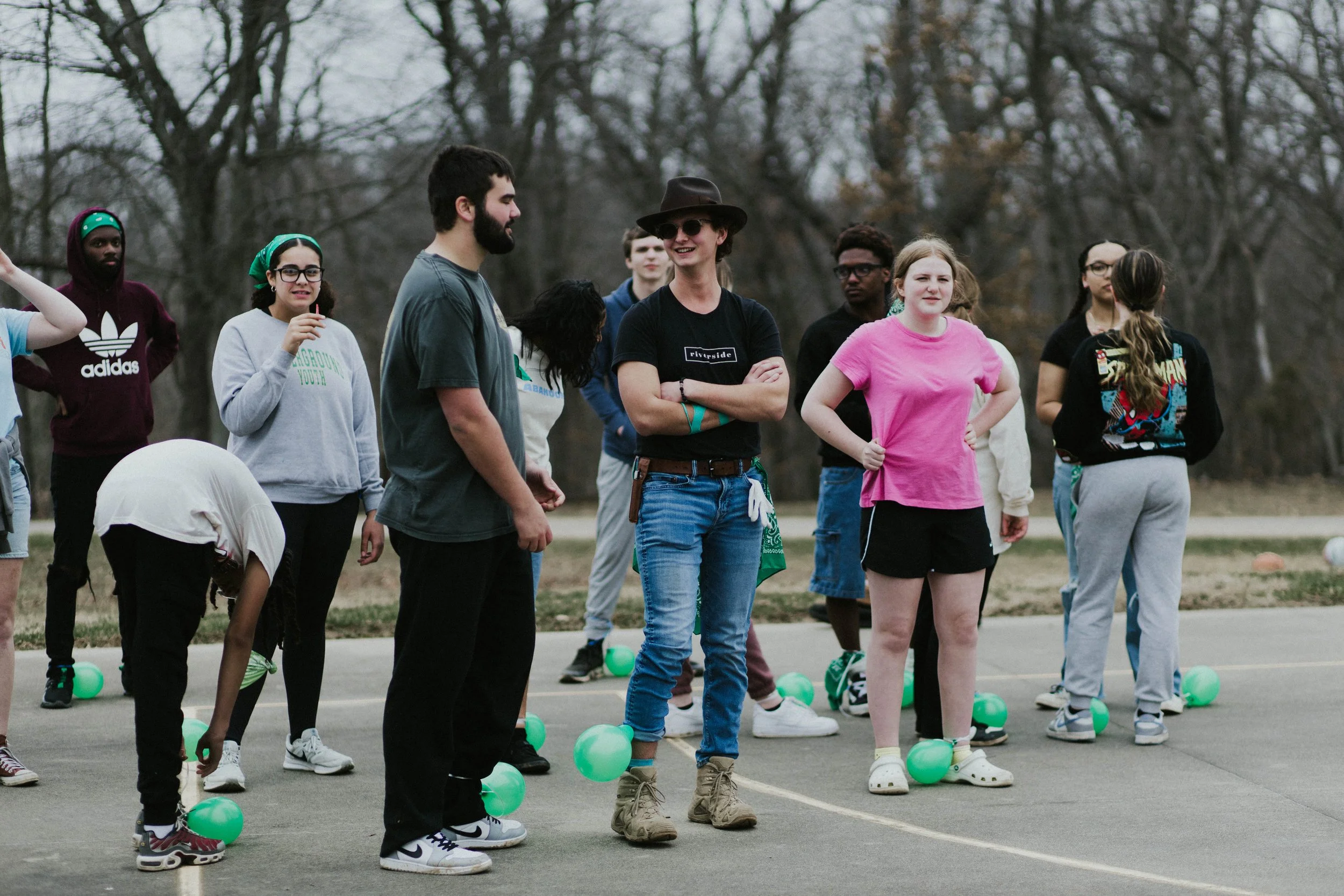 A group of young people standing outdoors on a court, some with green balloons, in a casual setting with trees in the background.
