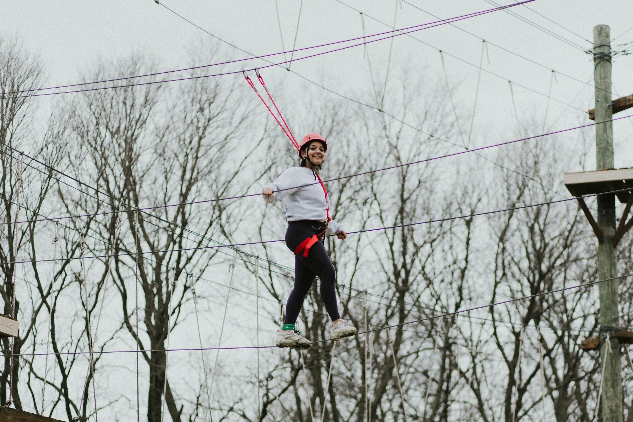 A young girl wearing a gray hoodie, black leggings, and a pink helmet walks on a ropes course outdoors, surrounded by leafless trees.