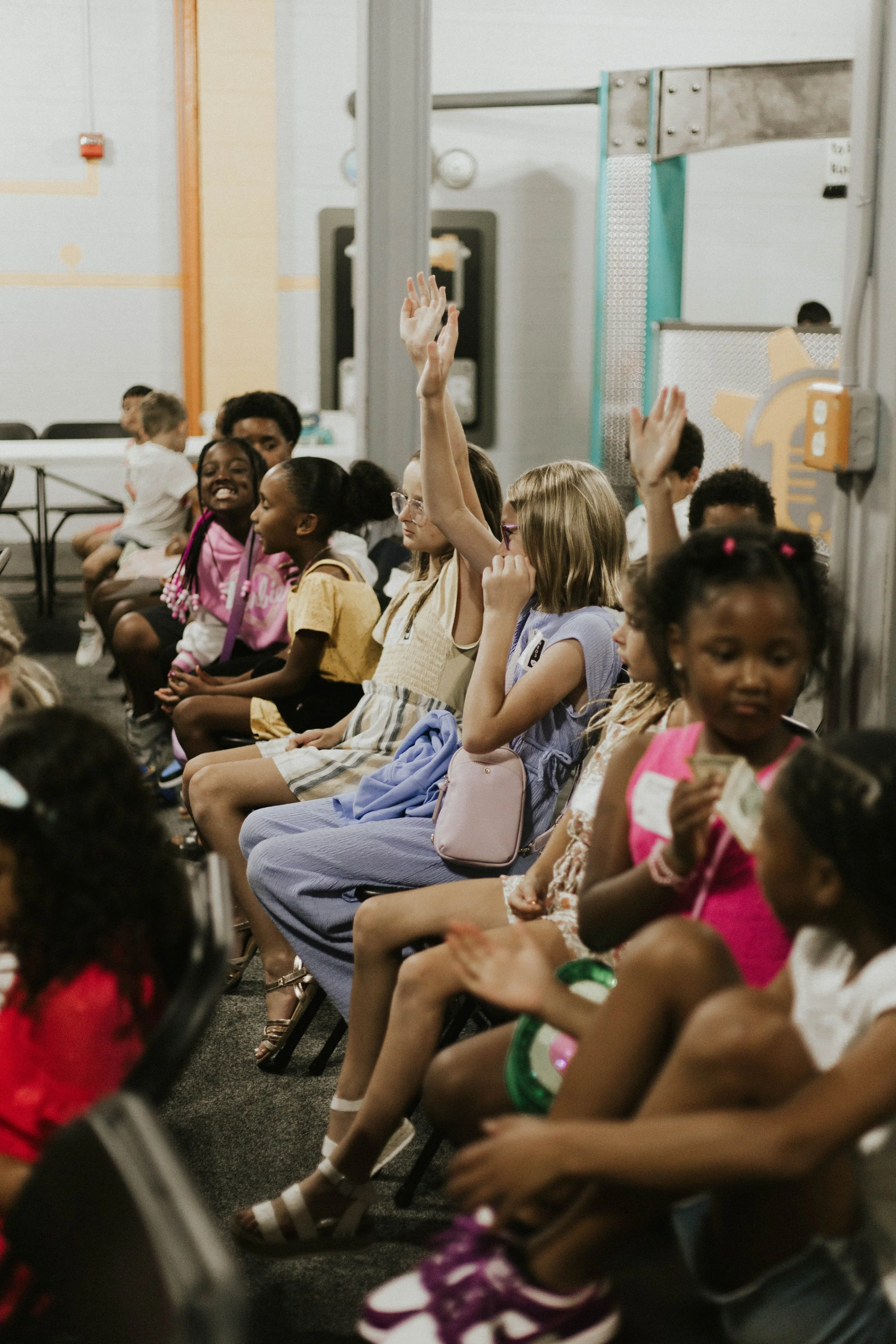 Children sitting in chairs during a classroom or assembly, with some raising their hands.