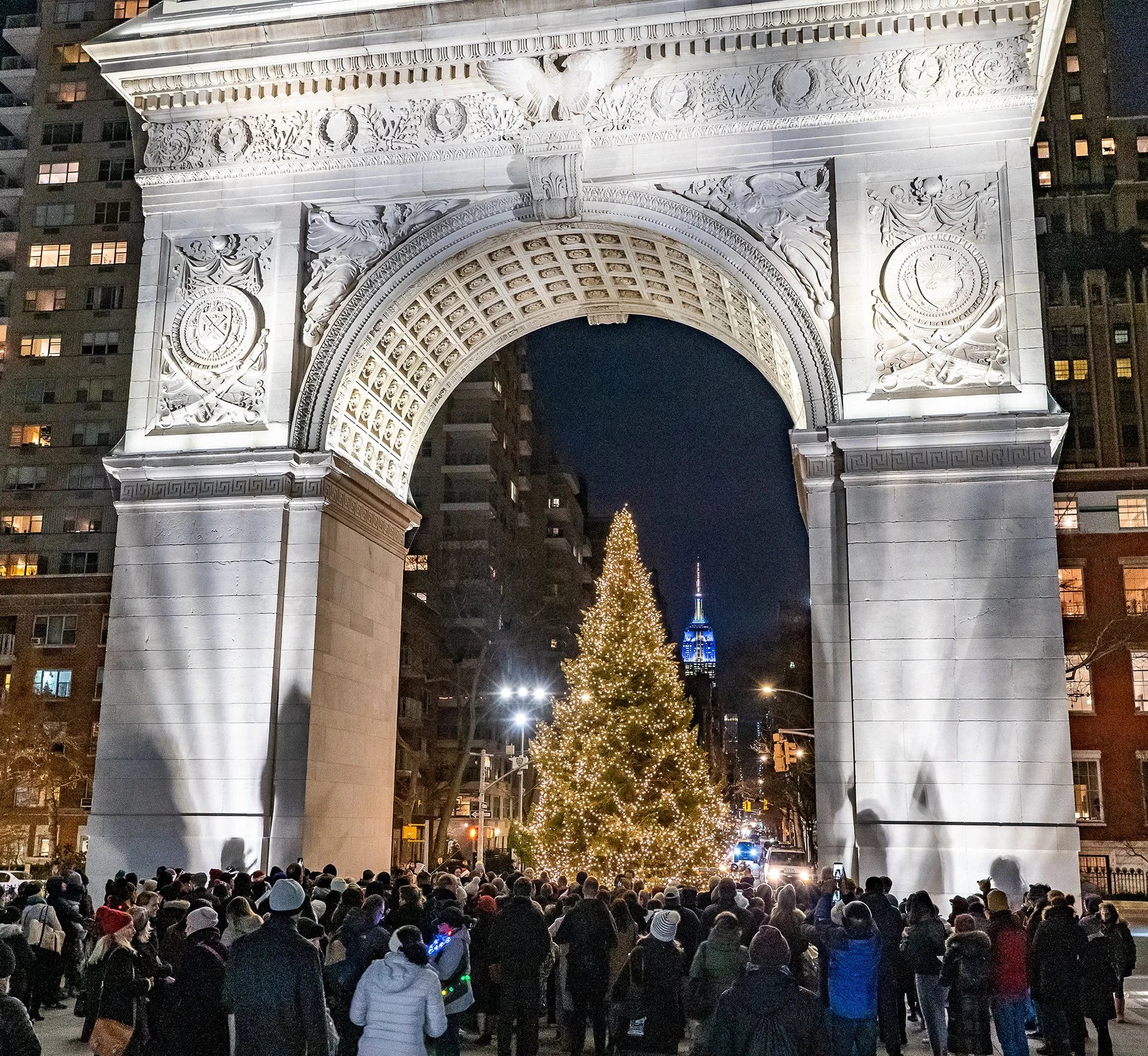 On Christmas Eve, a view of the warmly lit holiday tree framed by the Washington Square Arch and surrounded by carolers, the Empire State building is in the background lit in blue.