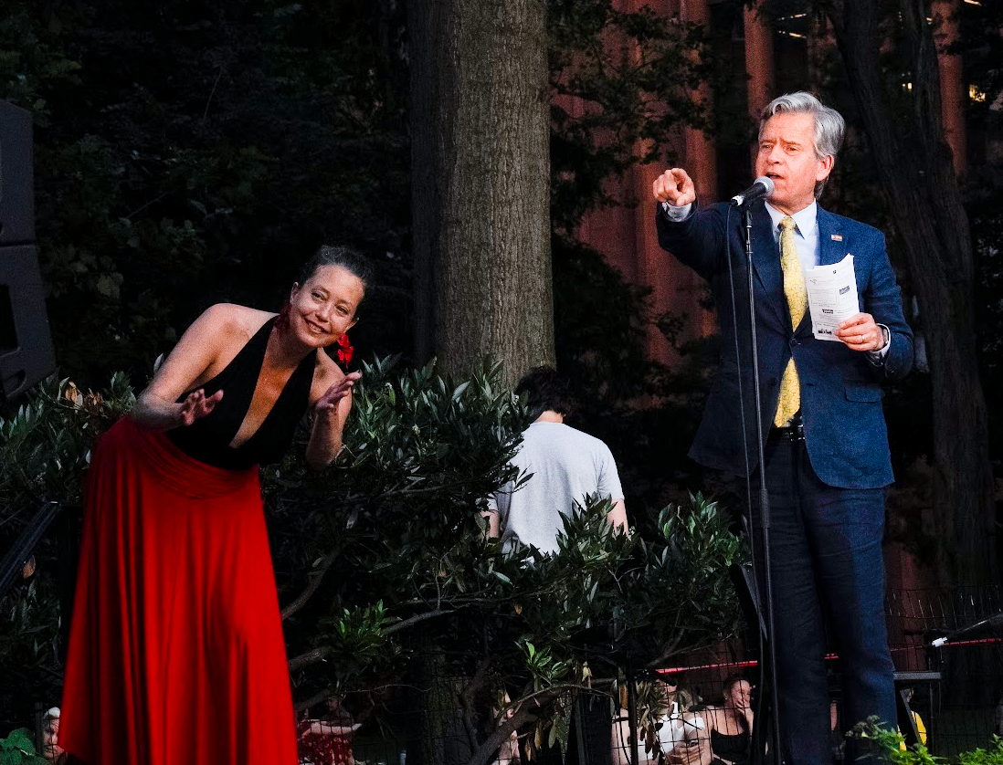 Manhattan Borough President, Brad Hoylman-Sigal, gives a jovial shout-out to CB2 Chair Valerie De La Rosa (off-screen) as Washington Square Association President, Erika Sumner (in the red skirt), peeks around speakers to applaud. Photo by Ken Howard.