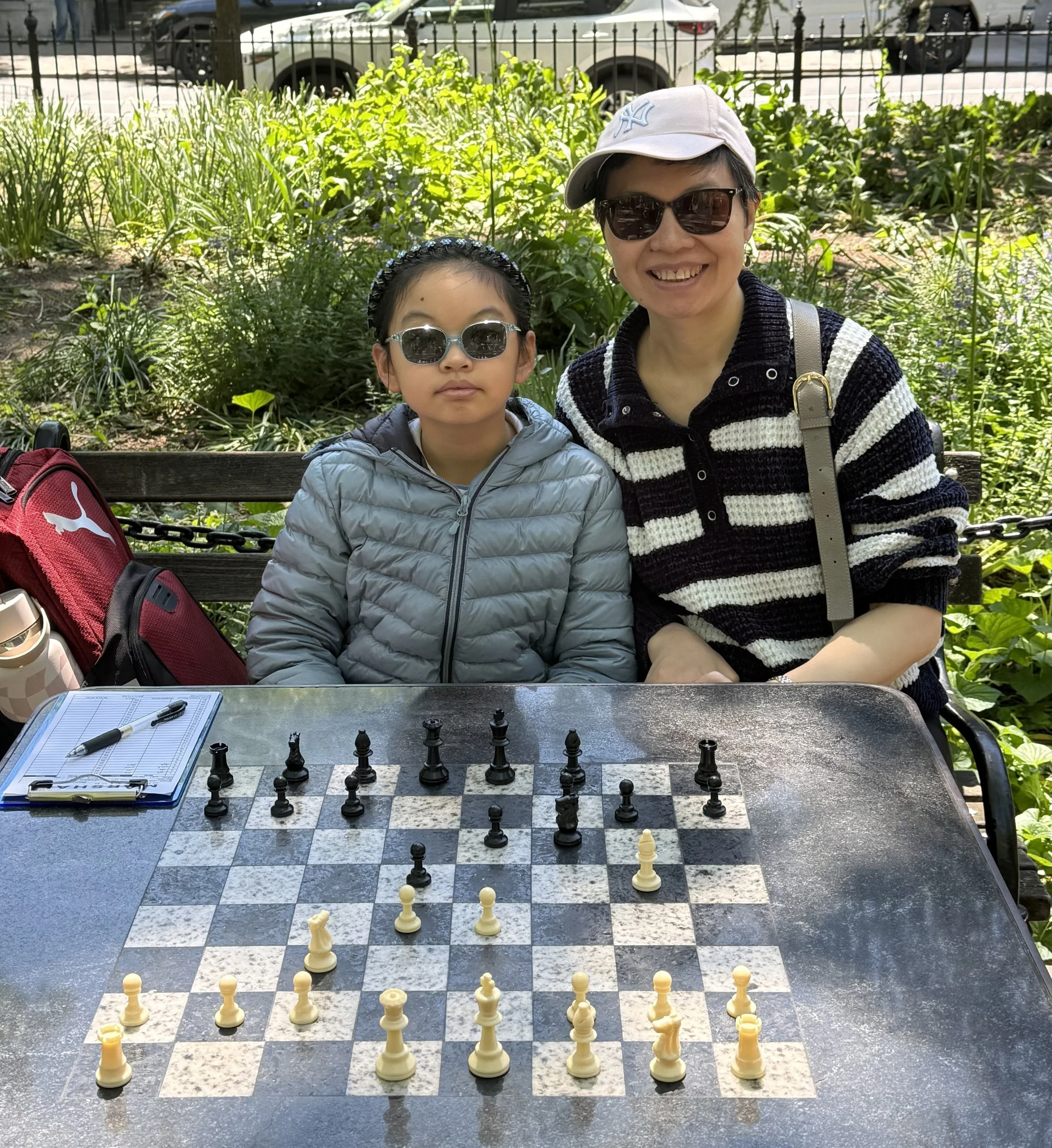 Abigail Zhou is seated at a chess table  wearing sunglasses next to Chunhua Weng
