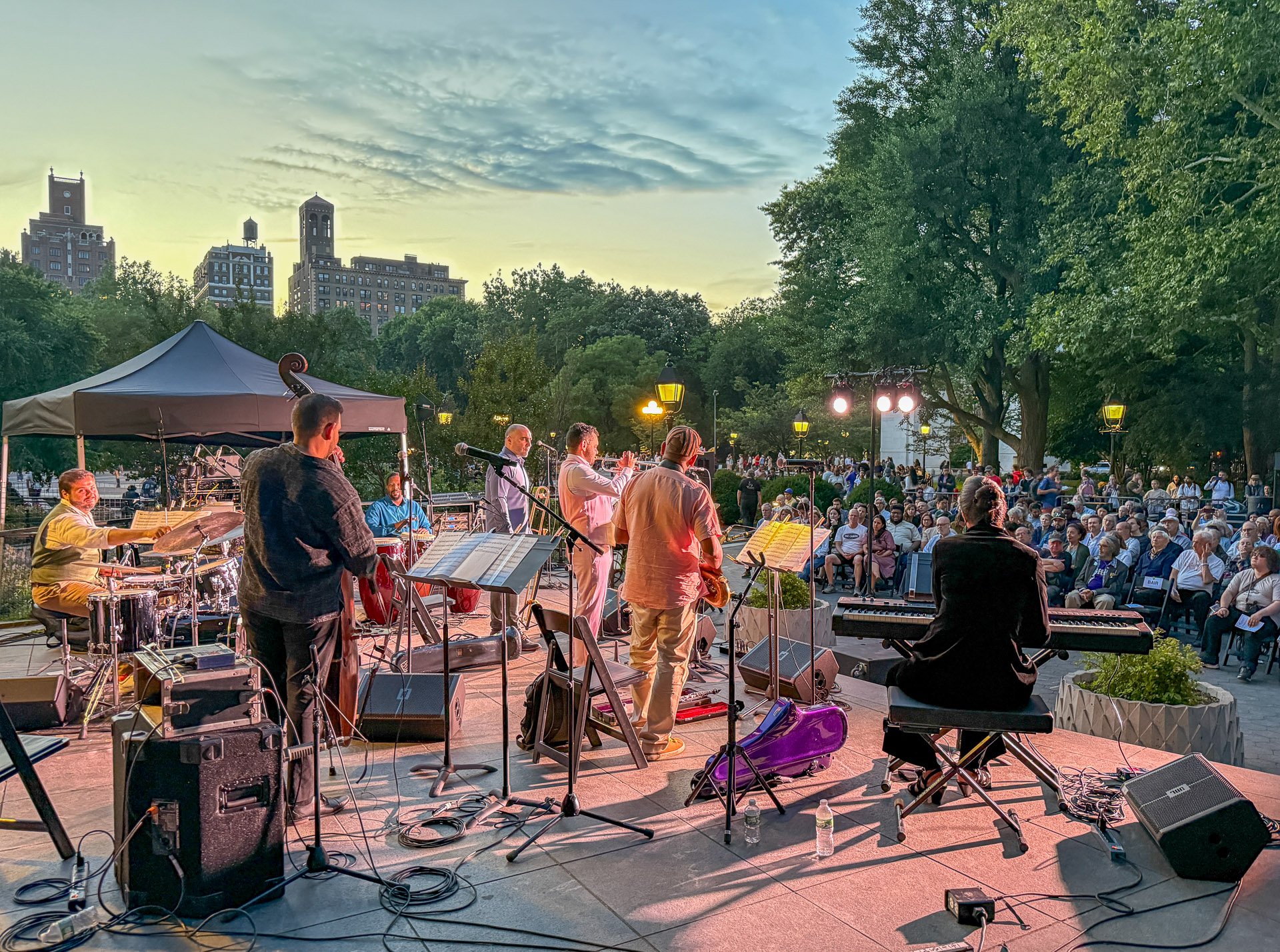 A view from behind Kali Rodriguez Peña and The New Cuban Masters Ensemble looking toward the audience with framed by the park’s west side skyline and a blue-green sunset