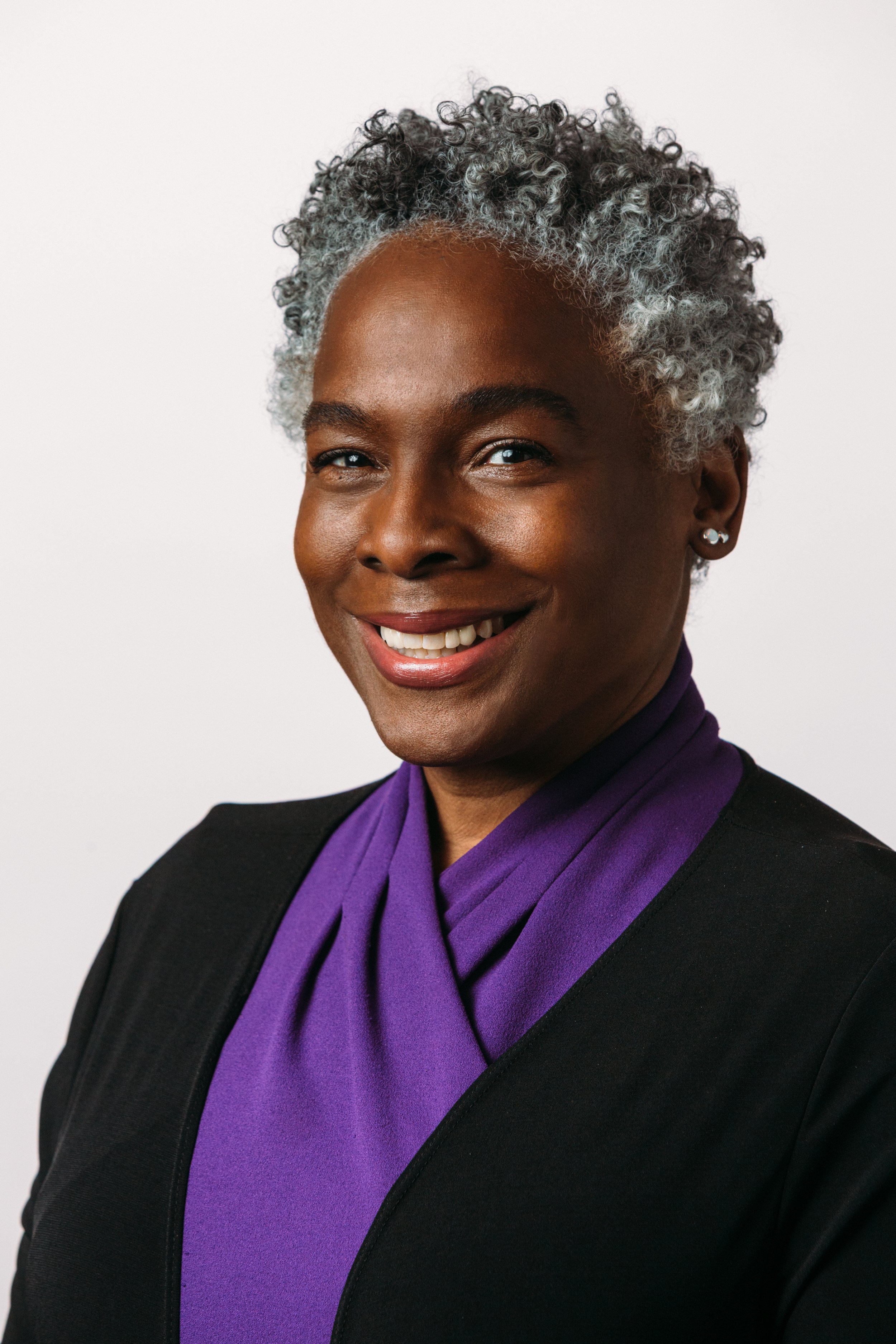 A Black woman with short curly peppered hair, wearing a purple shirt with black cardigan, smiling at the camera.