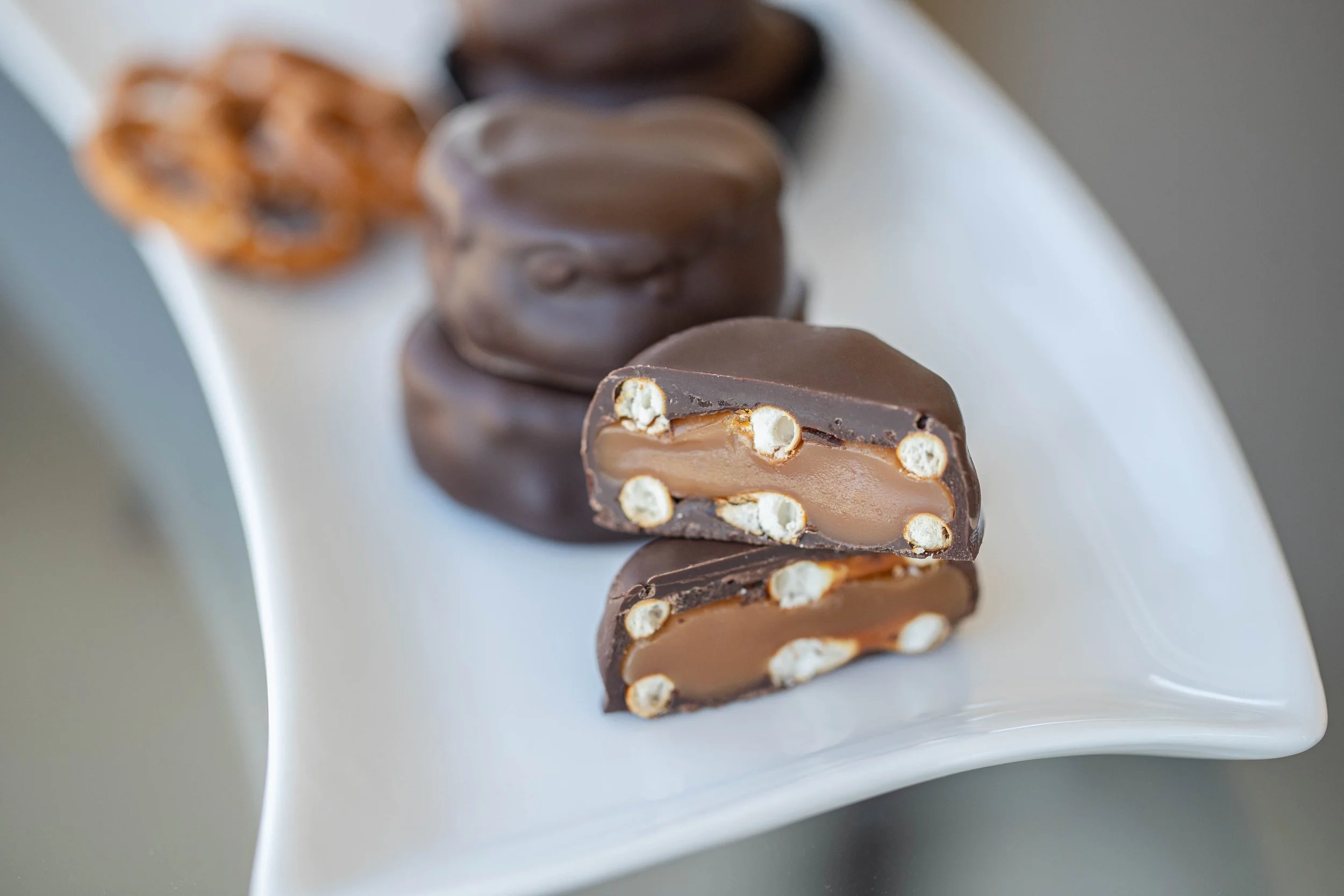 Close-up of a layered chocolate and caramel candy bar with marshmallow pieces, sliced in half to reveal the filling, placed on a white rectangular plate.