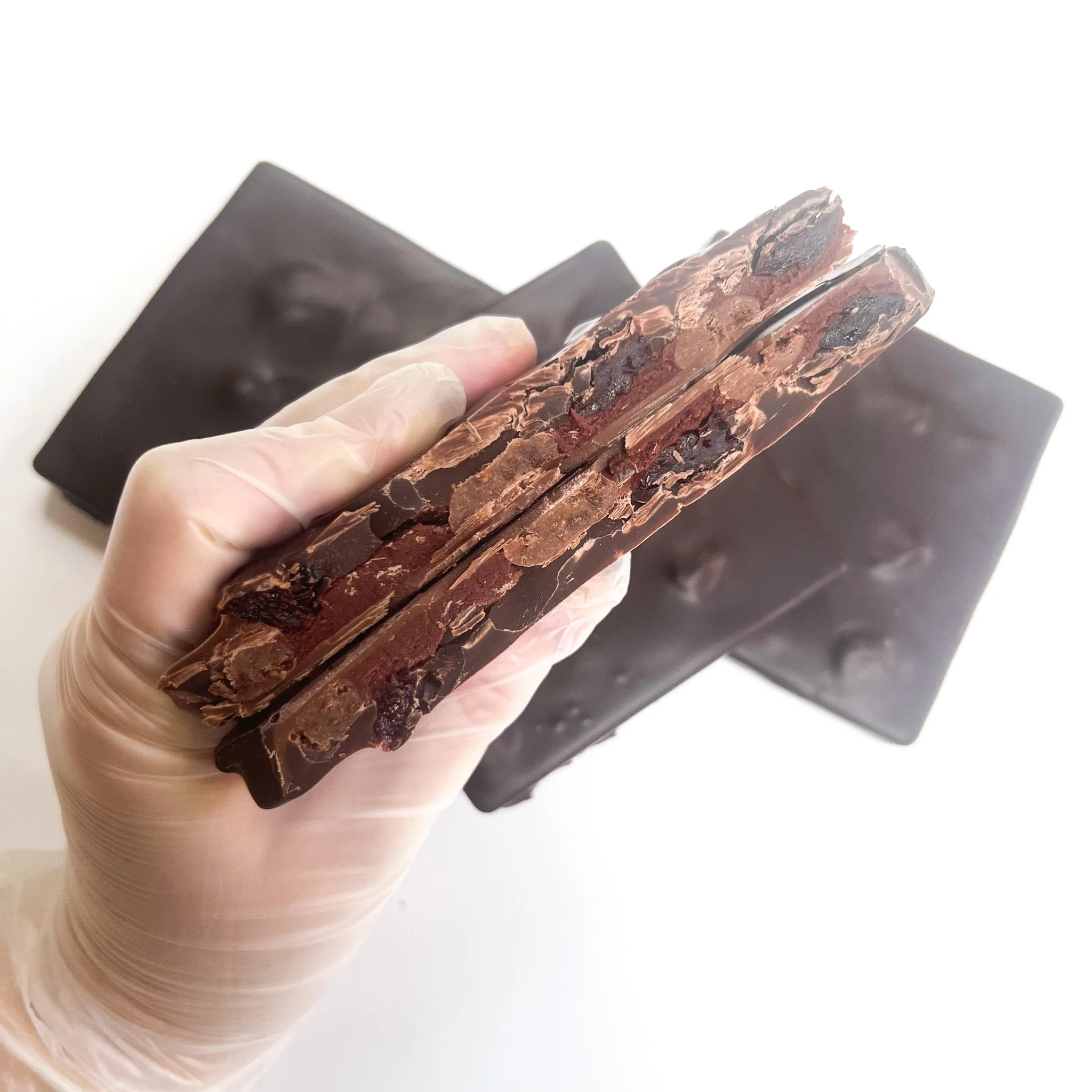 Close-up of a person's hand holding a broken piece of chocolate with dark, melted chocolate and nuts inside, with a black object in the background.