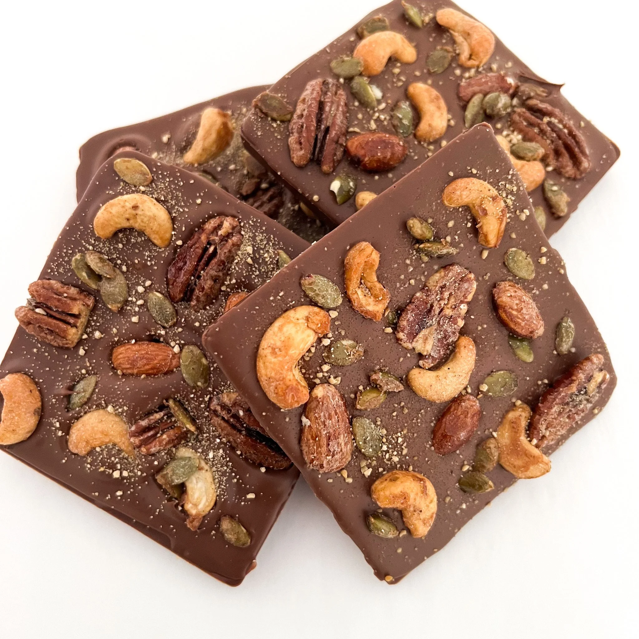 Close-up of two pieces of dark chocolate topped with mixed nuts, including cashews, almonds, pecans, and pumpkin seeds, on a white background.