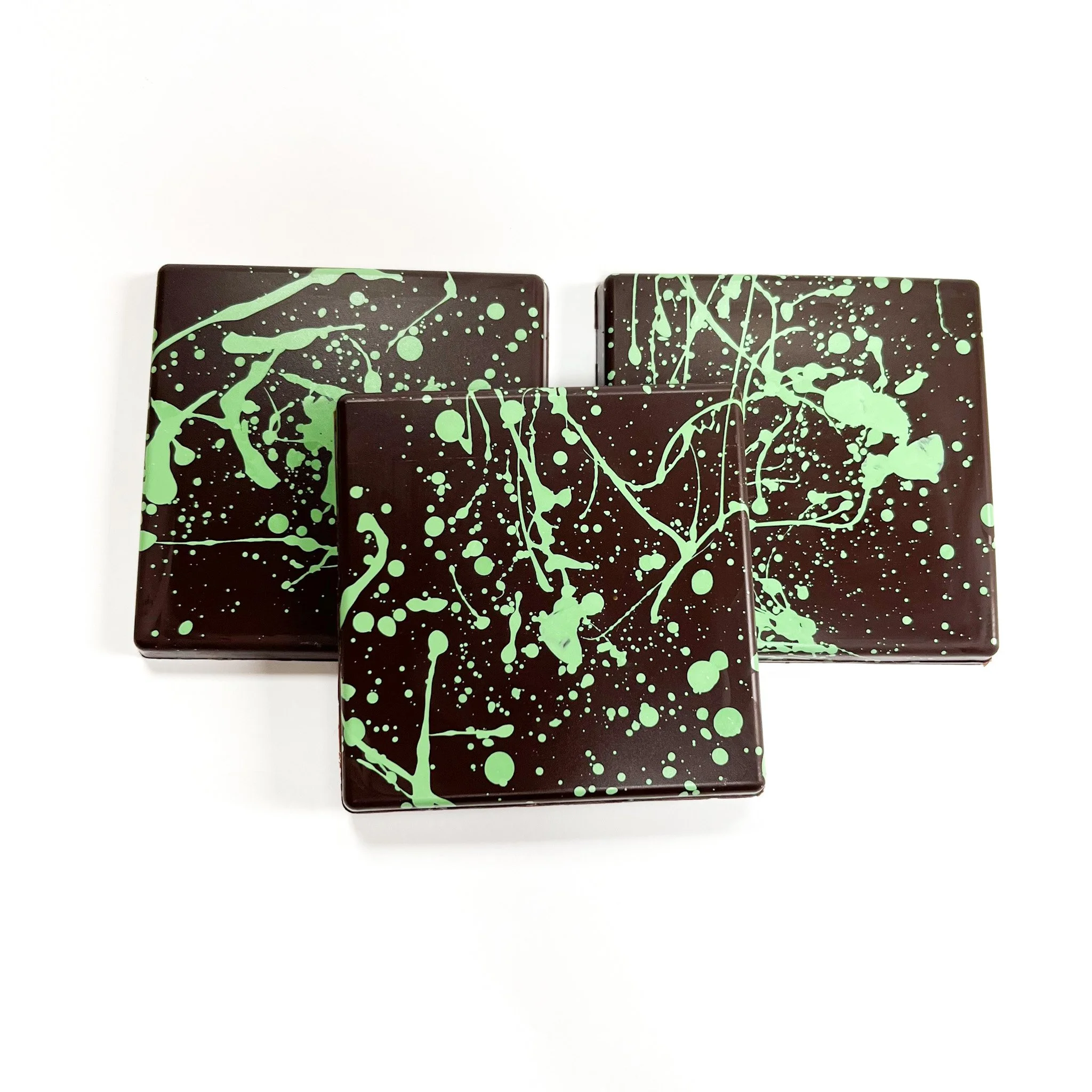 Close-up of three rectangular bars of dark chocolate with light green splatter patterns on top, arranged in a staggered manner against a white background.