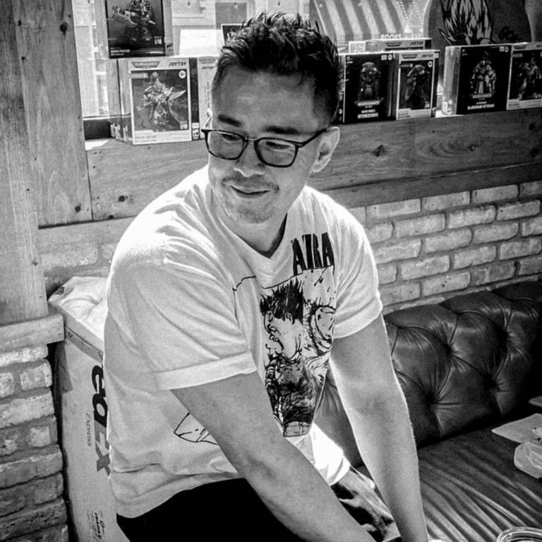 A man with glasses and short dark hair sitting on a leather couch in a comic book or figure display store, wearing a white T-shirt with a graphic. Behind him, toys or action figures are displayed on a wooden shelf, with a brick wall in the background.