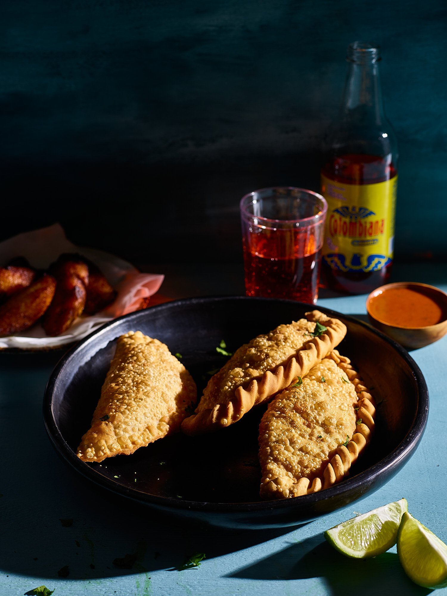 A black plate with three fried empanadas garnished with herbs, a side of lime wedges, a small bowl of dipping sauce, a glass of red soda, a bottle of soda, and a plate of chicken wings in the background.