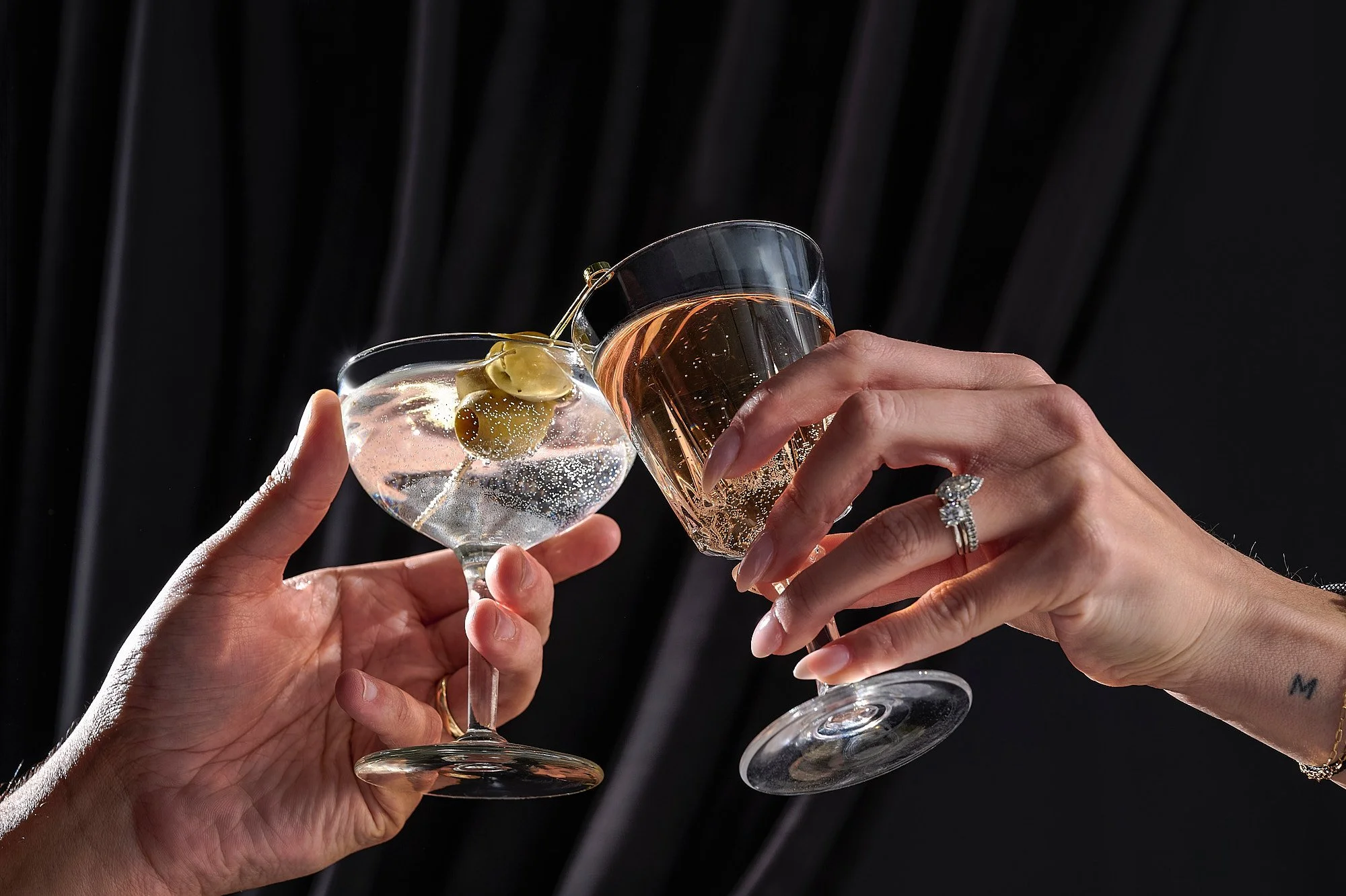 Two hands holding glasses of champagne and a cocktail toast in a celebratory gesture.
