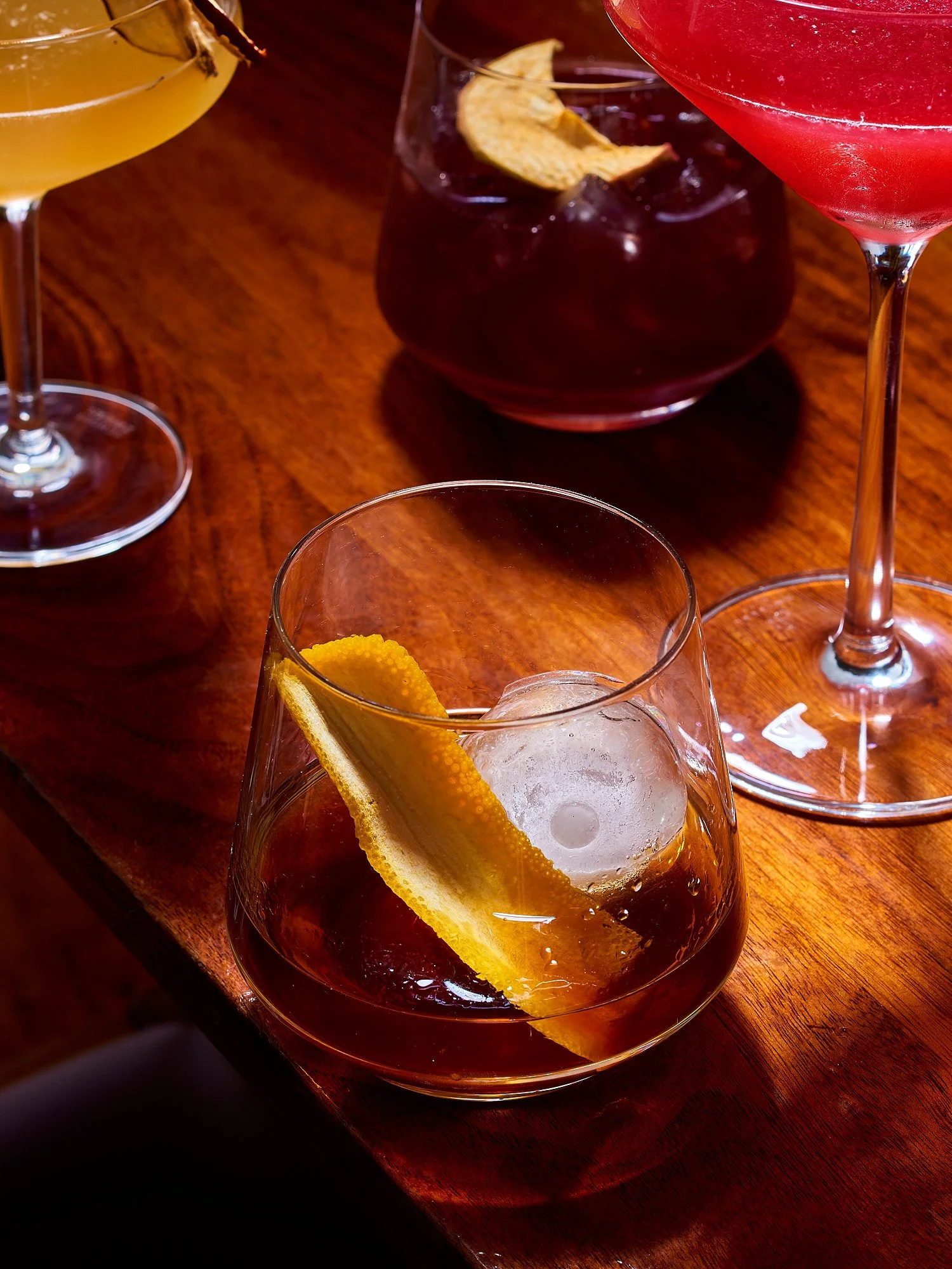 Spirits advertising photography by Dhanraj Emanuel — whiskey cocktail with orange peel and large ice cube on wooden bar surface. Commercial beverage photographer for spirits brands and advertising agencies, working nationally.