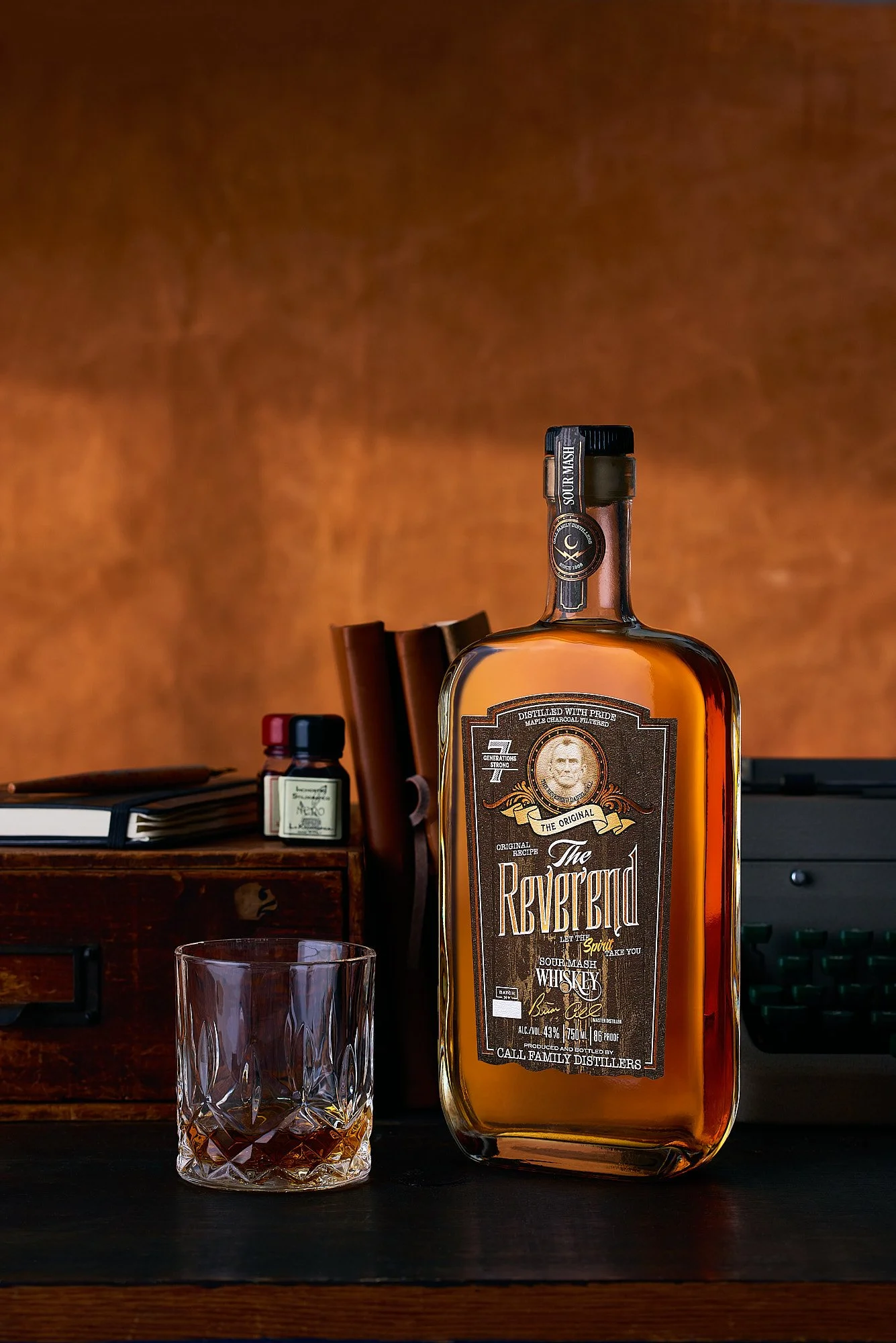 Spirits advertising photography by Dhanraj Emanuel — The Reverend whiskey bottle styled with glass and wooden box on dark surface, warm dramatic lighting. Commercial beverage photographer for craft spirits brands, available nationwide.