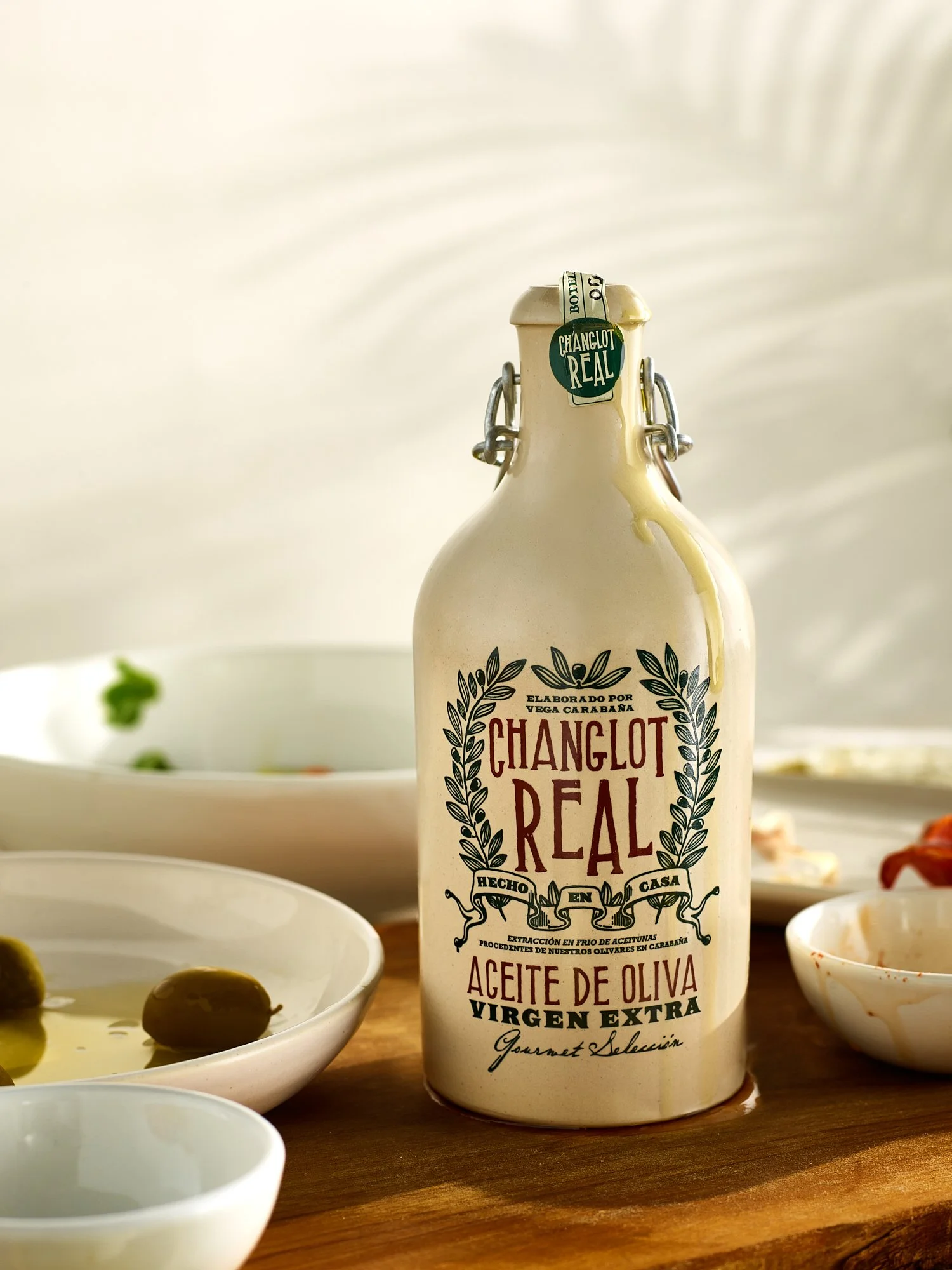  Changlot Real extra virgin olive oil in its stoneware swing-top bottle standing on a wooden board among olives, dishes, and an antipasto spread, backlit with soft window light. Food and beverage advertising photography by Dhanraj Emanuel. 