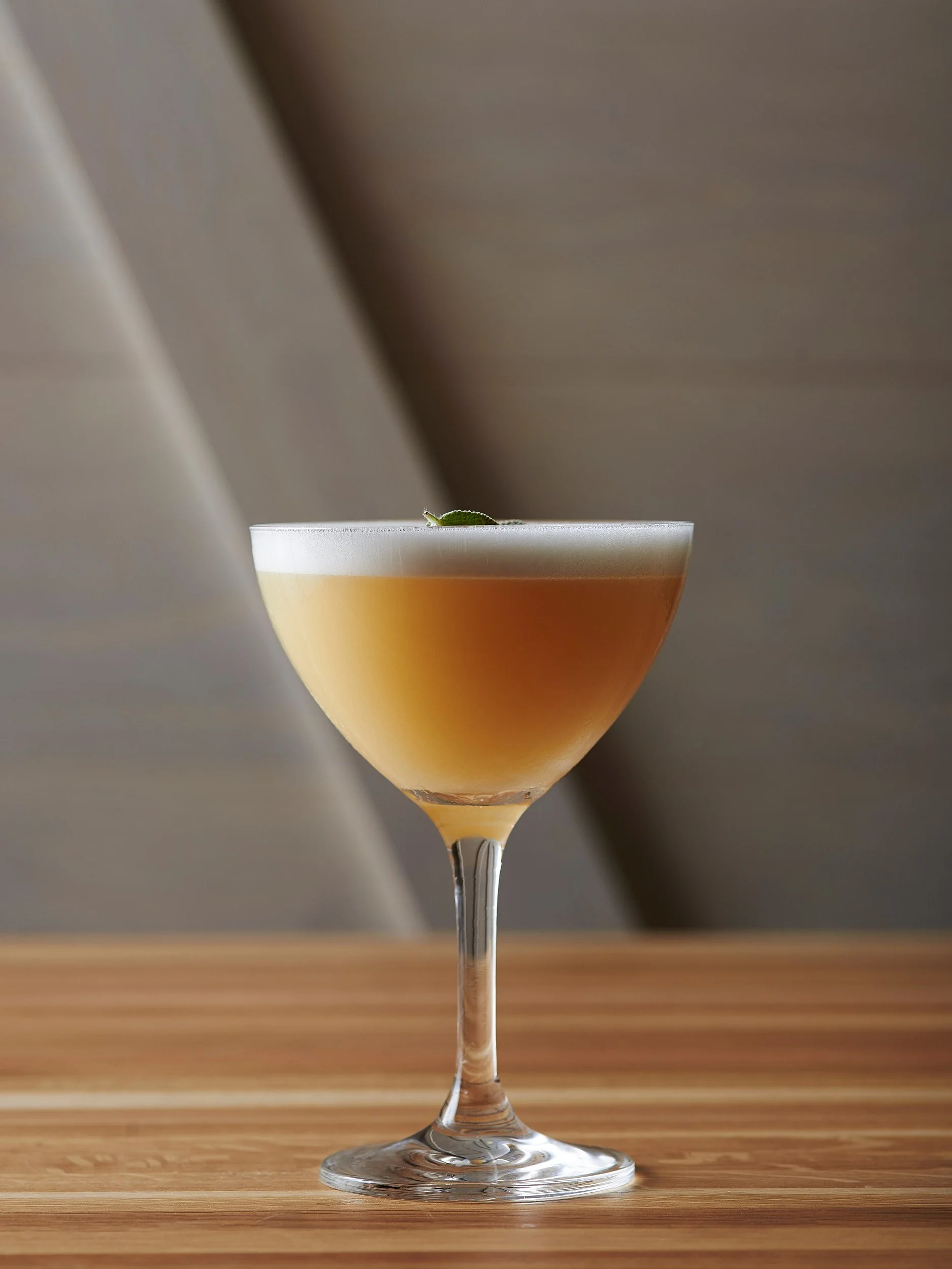A cocktail in a coupe glass with a frothy top and a small garnish on the rim, set on a wooden surface.